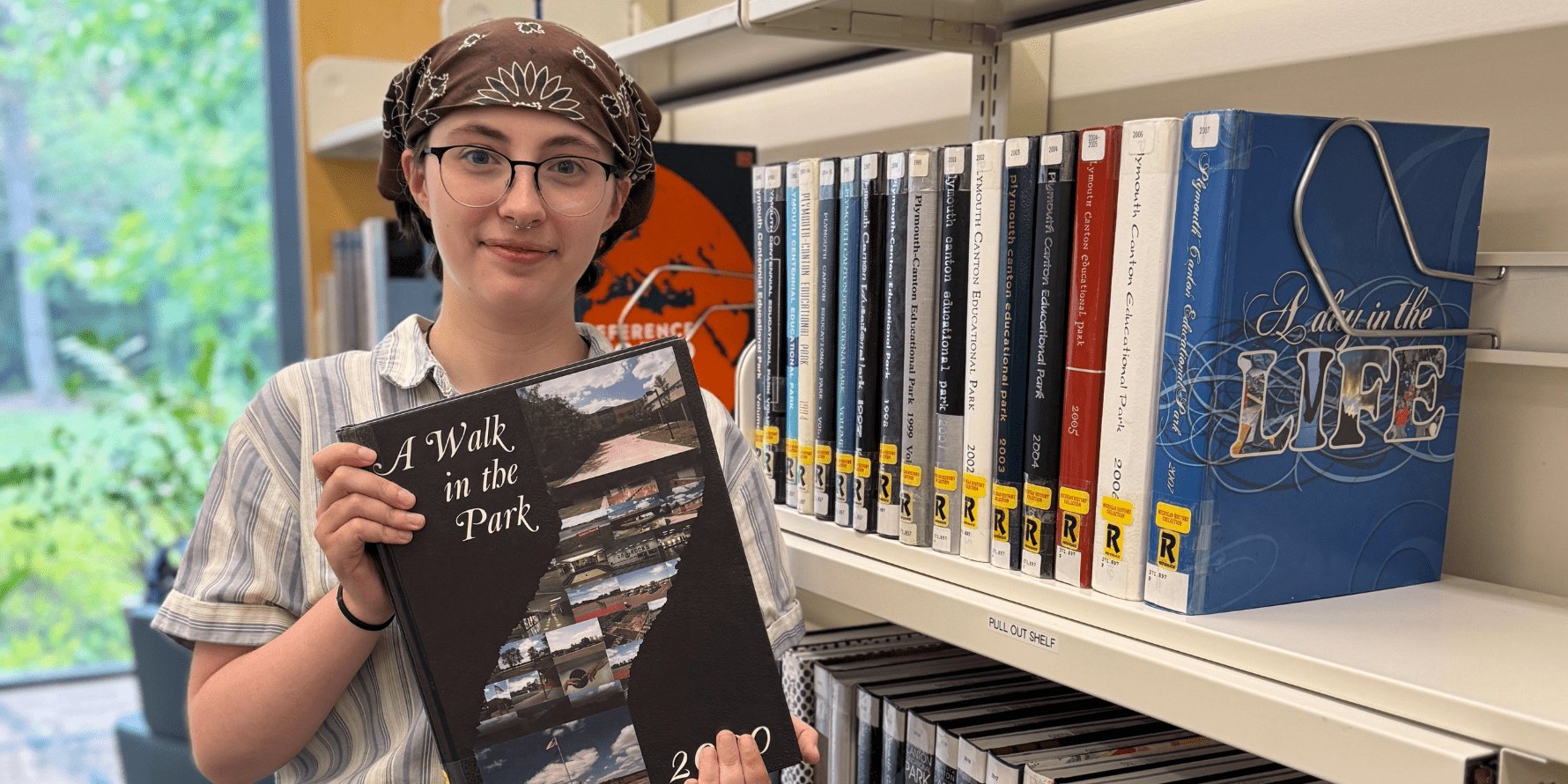 Person holding a school yearbook stands beside a library shelf of books.