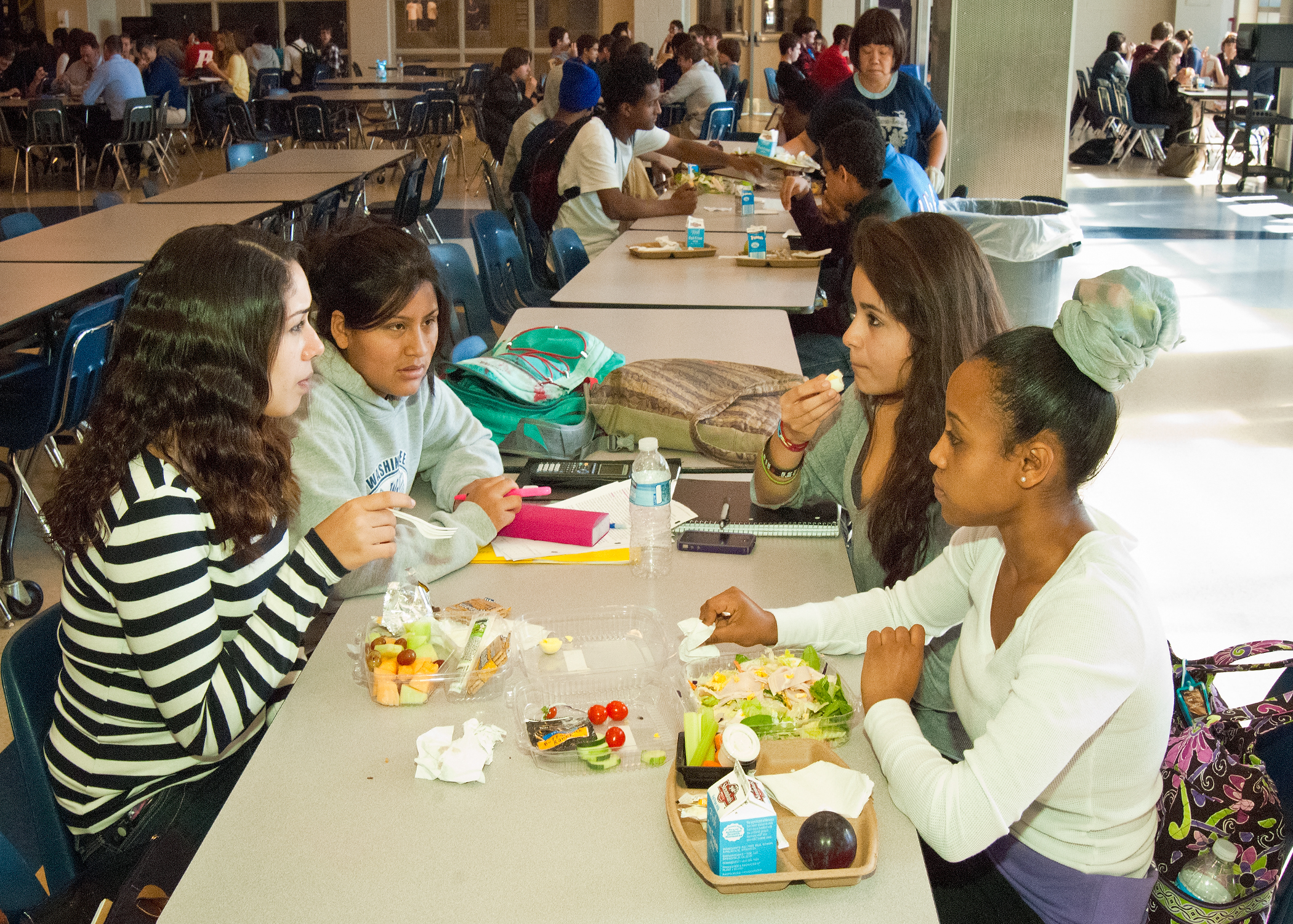Students eat salads and fruit together at tables in a school cafeteria.