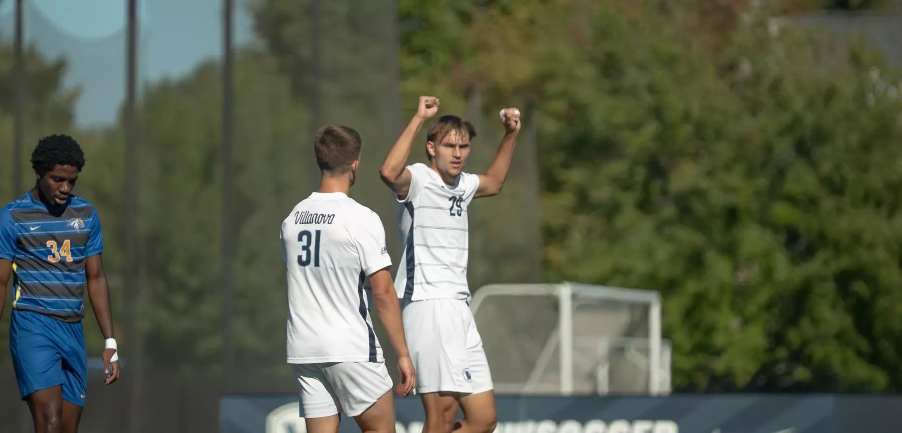 Soccer player in white uniform raises both fists in celebration on a grass field.