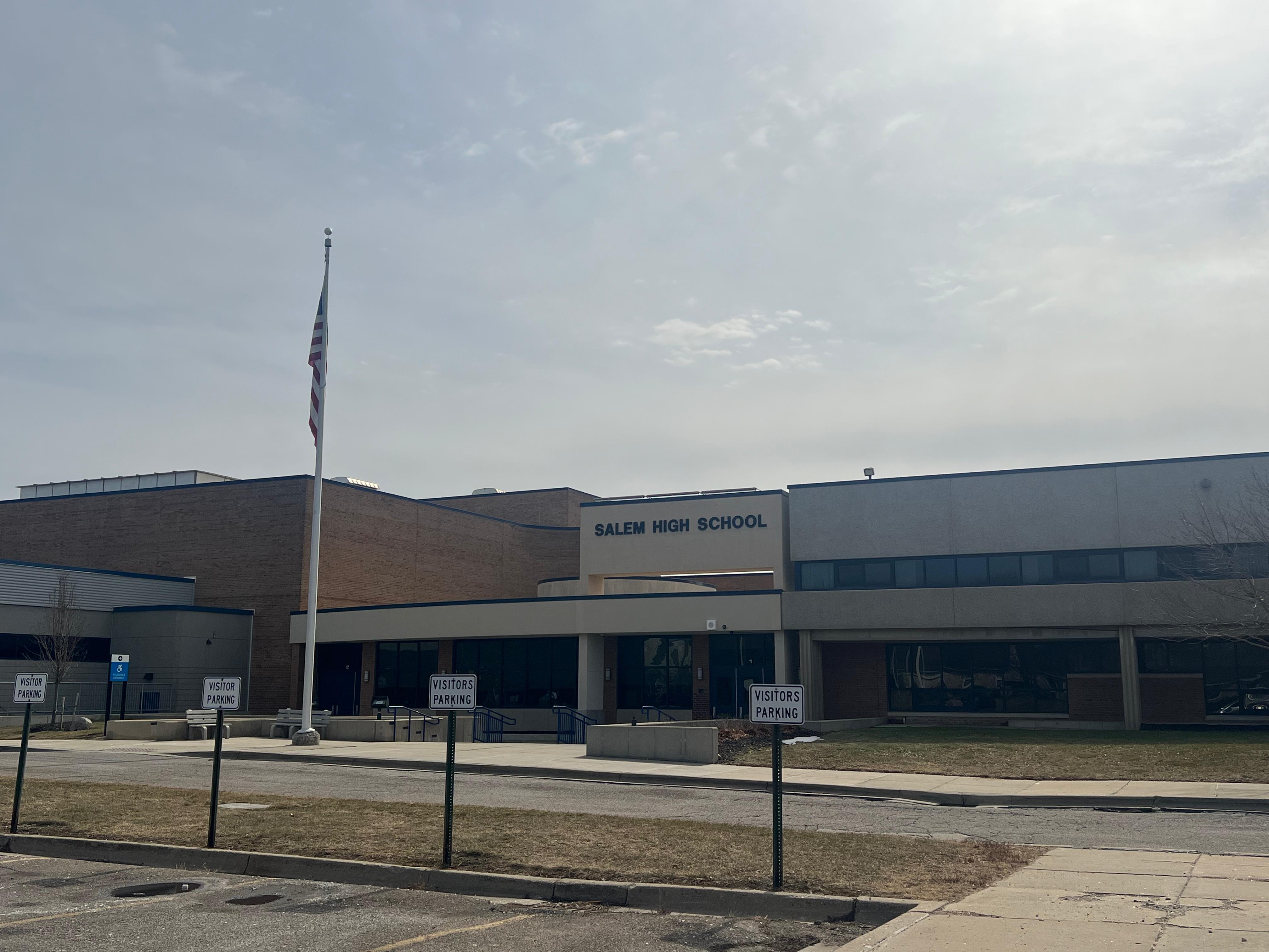Brick school building exterior with flagpole and visitor parking signs under an overcast sky.