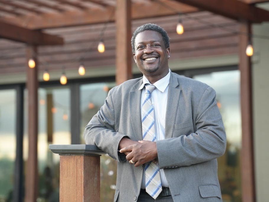A smiling man in a gray suit and striped tie leaning on a wooden post in an outdoor patio setting.