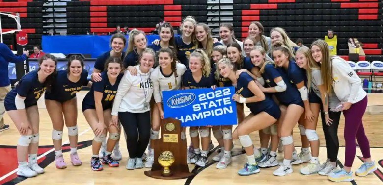 Female volleyball players in navy and gold uniforms pose with a state championship trophy in a gymnasium.