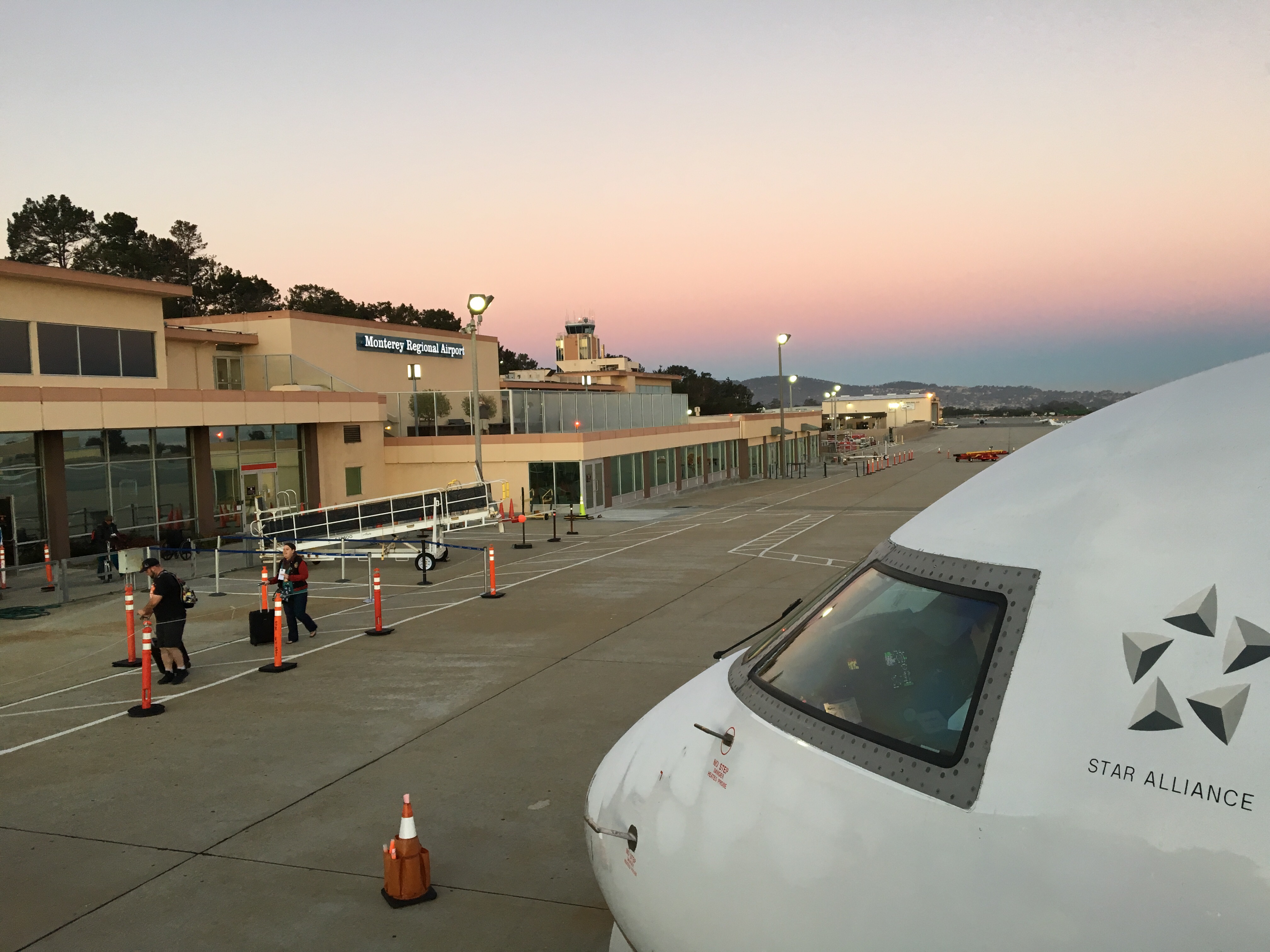 Passengers walk across an airport tarmac toward a parked aircraft at sunrise.