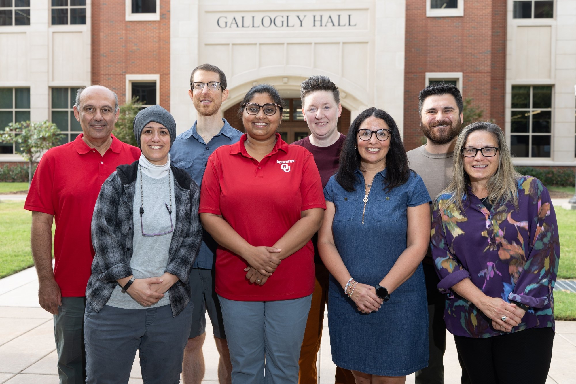 Group of eight adults smiling together in front of a brick university building.