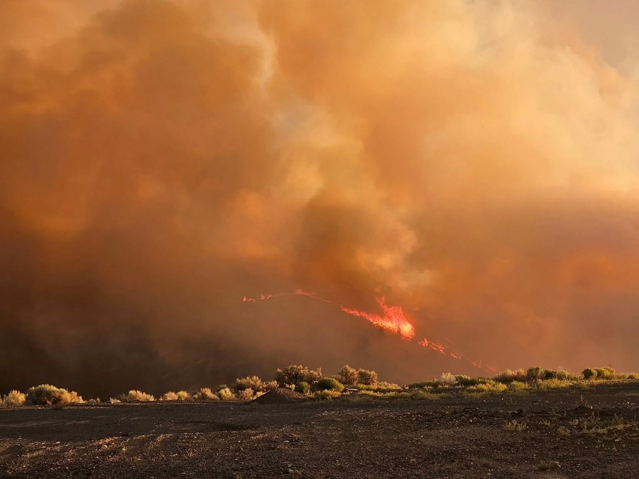 Large orange-brown smoke plumes rise from a burning ridge with charred ground in the foreground.