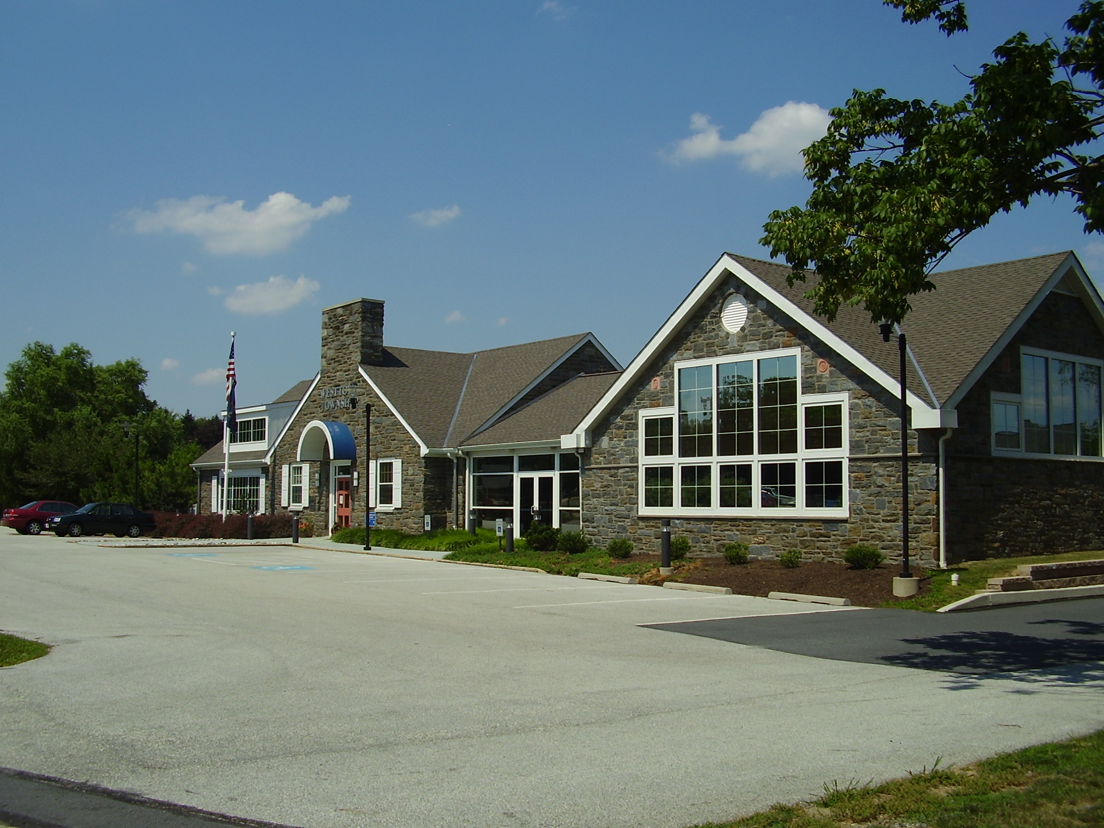 Stone and siding civic building with a flagpole and parking lot under a blue sky.