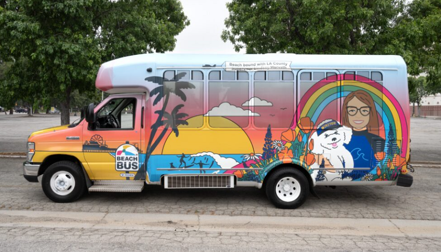 A small colorful shuttle bus labeled Beach Bus with artwork featuring a sunset, palm trees, rainbow, ocean waves, and illustrations of a woman and a dog.