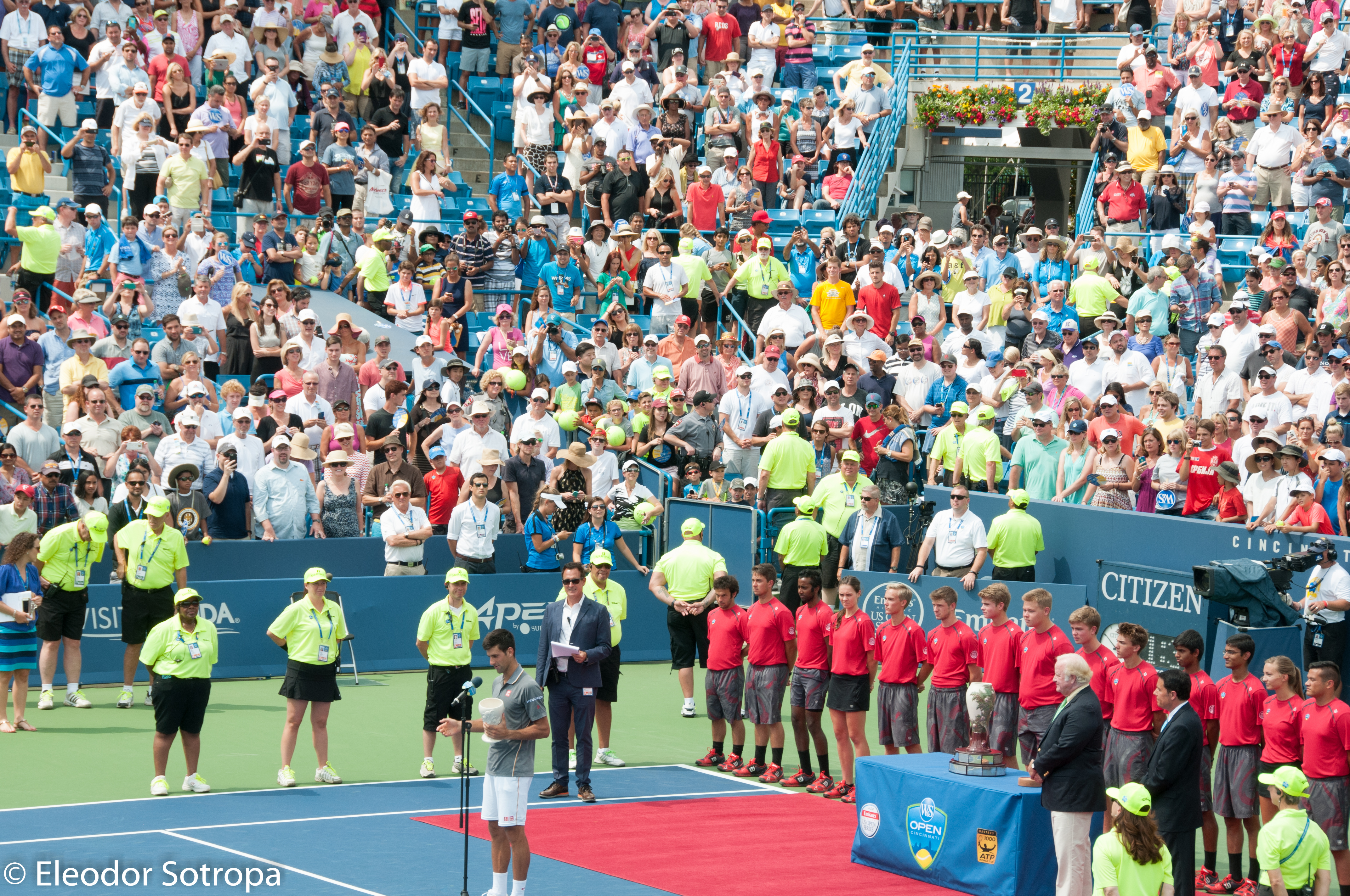 Participants lined up near a podium on a blue tennis court in a packed outdoor stadium.