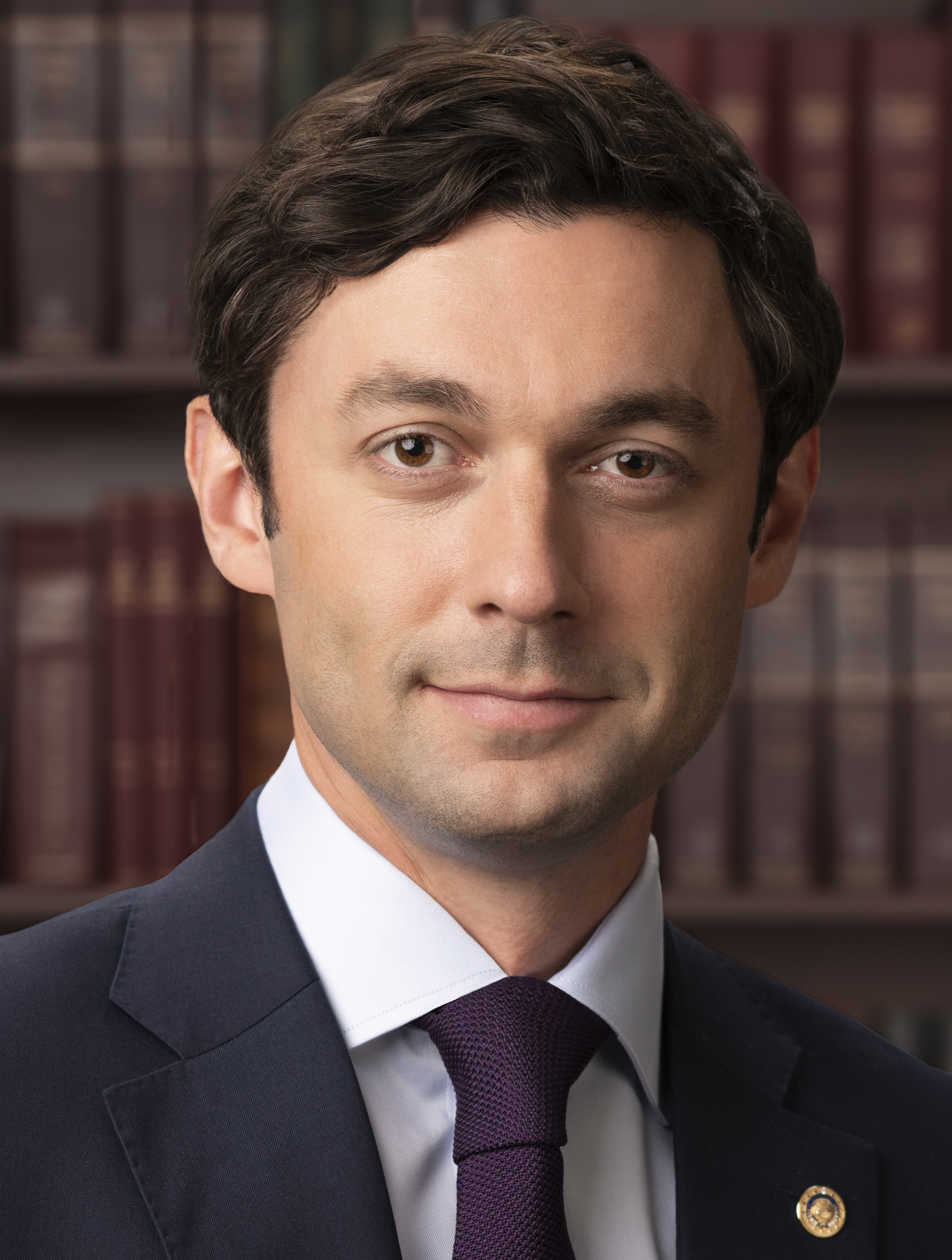 Man in dark suit and purple tie seated before a bookshelf in a formal portrait.