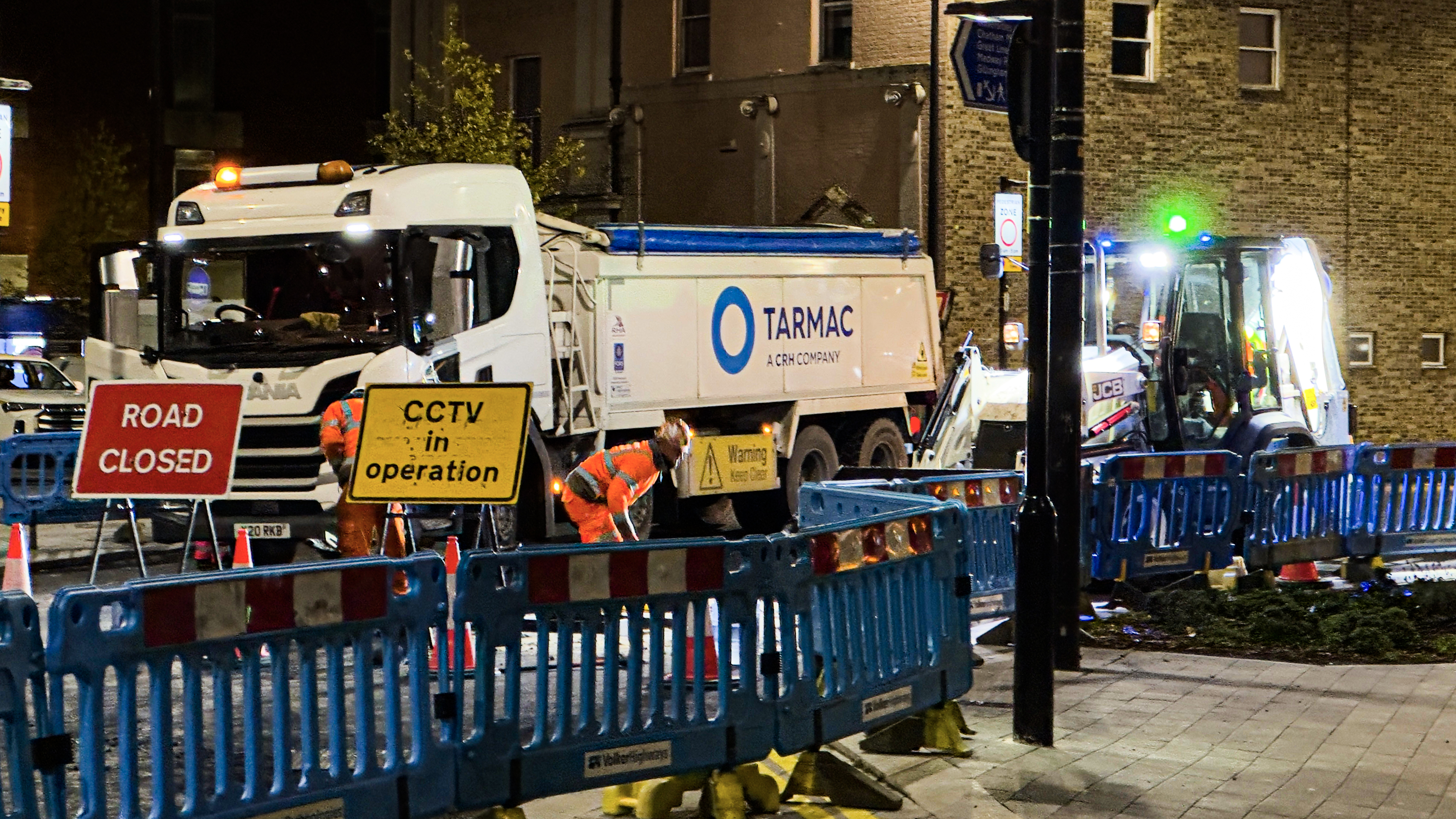 Workers in orange vests operate heavy equipment on a closed road at night.