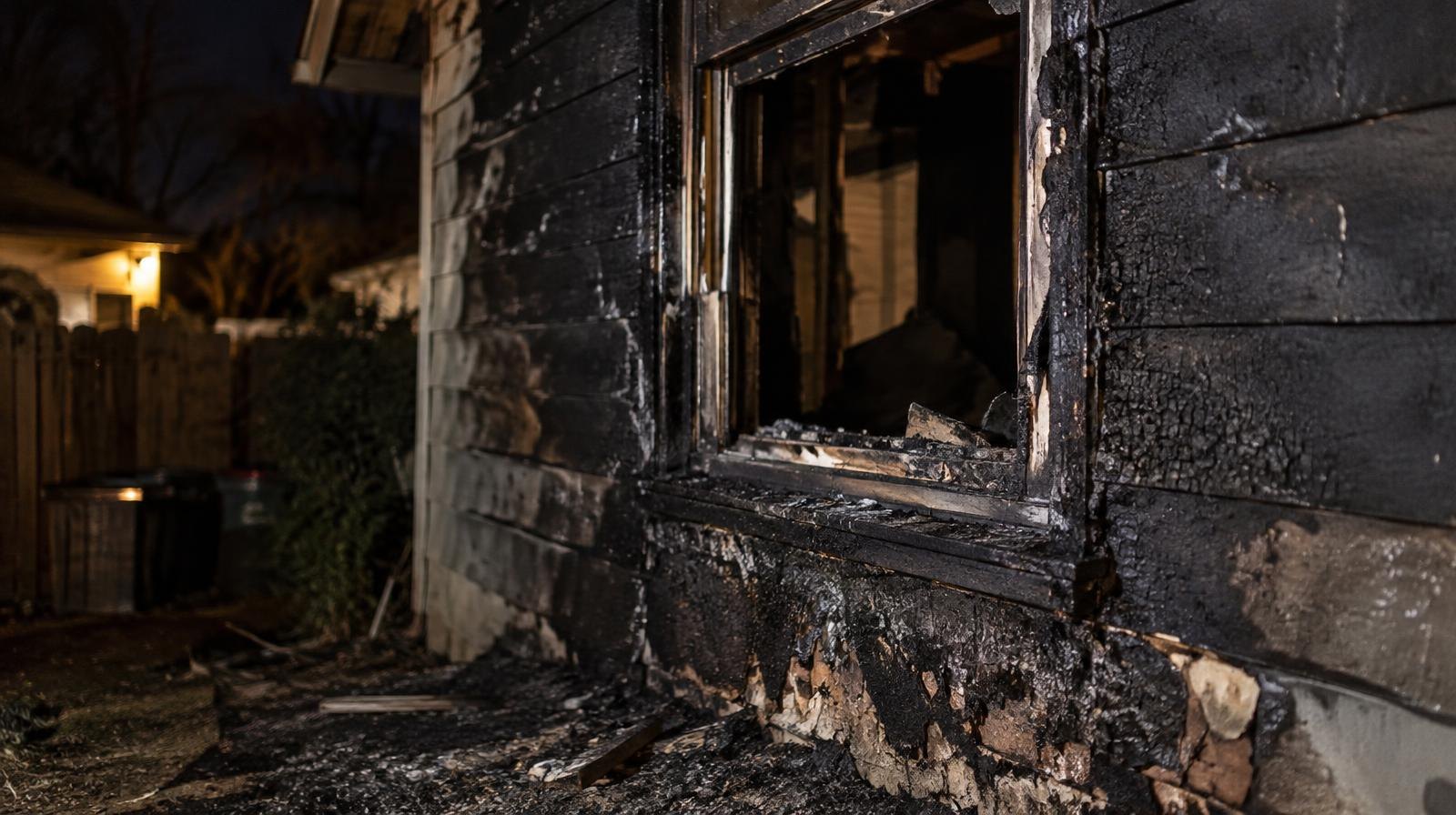 Smoke-blackened exterior wall of a small outbuilding with scorch marks around a window.