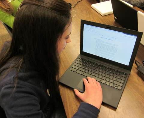 A student looks down at a Chromebook laptop on a wooden school desk.