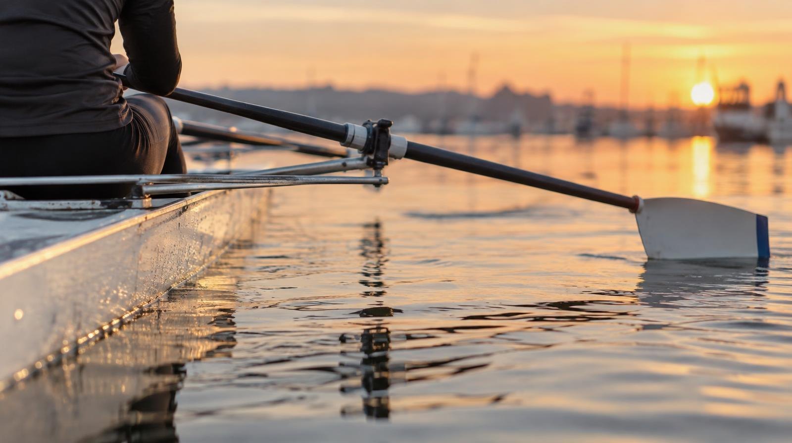 A lightweight rowing shell with oars extended over still water in early morning light.