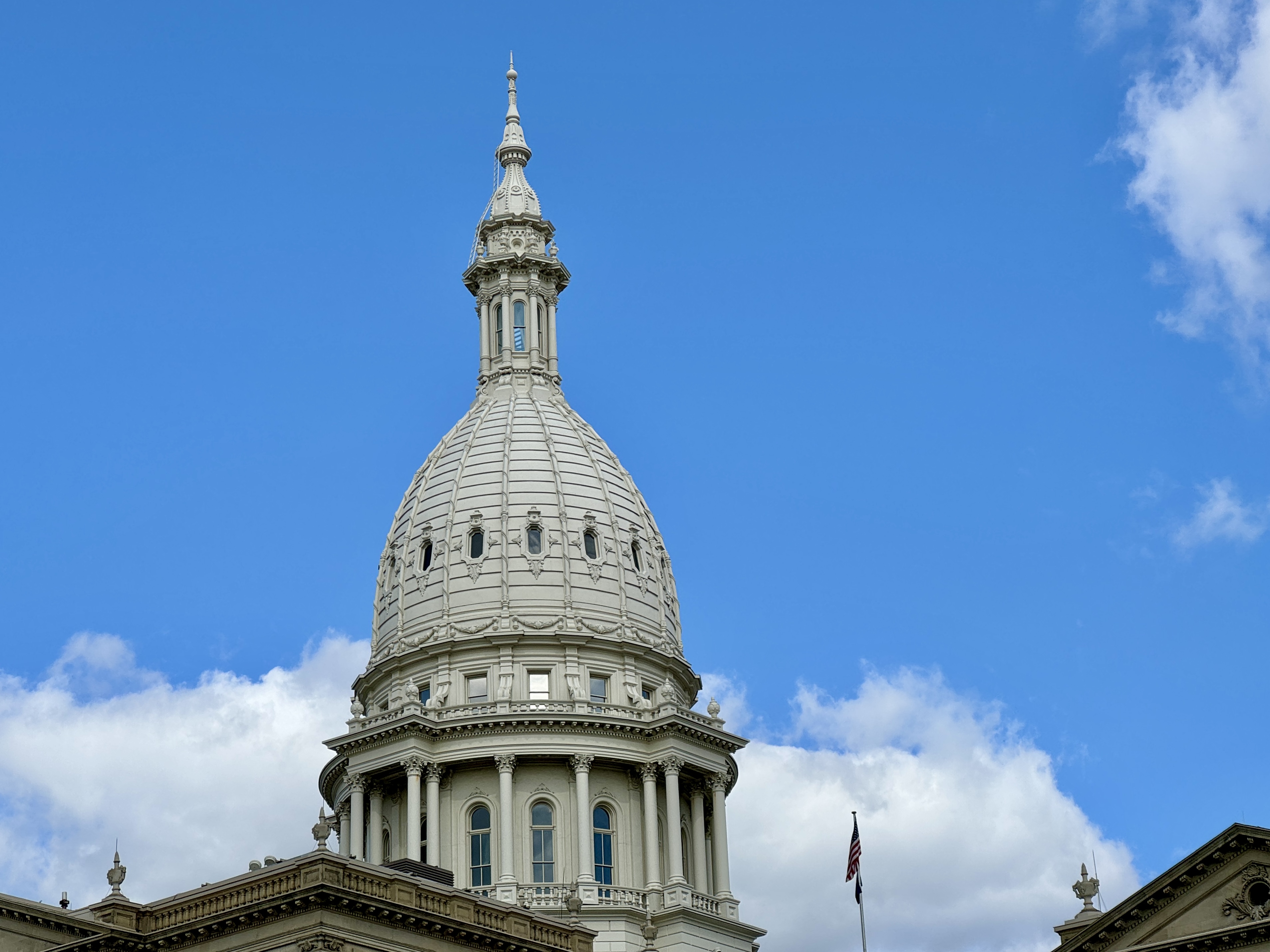Michigan State Capitol dome against a blue sky with scattered clouds and an American flag.