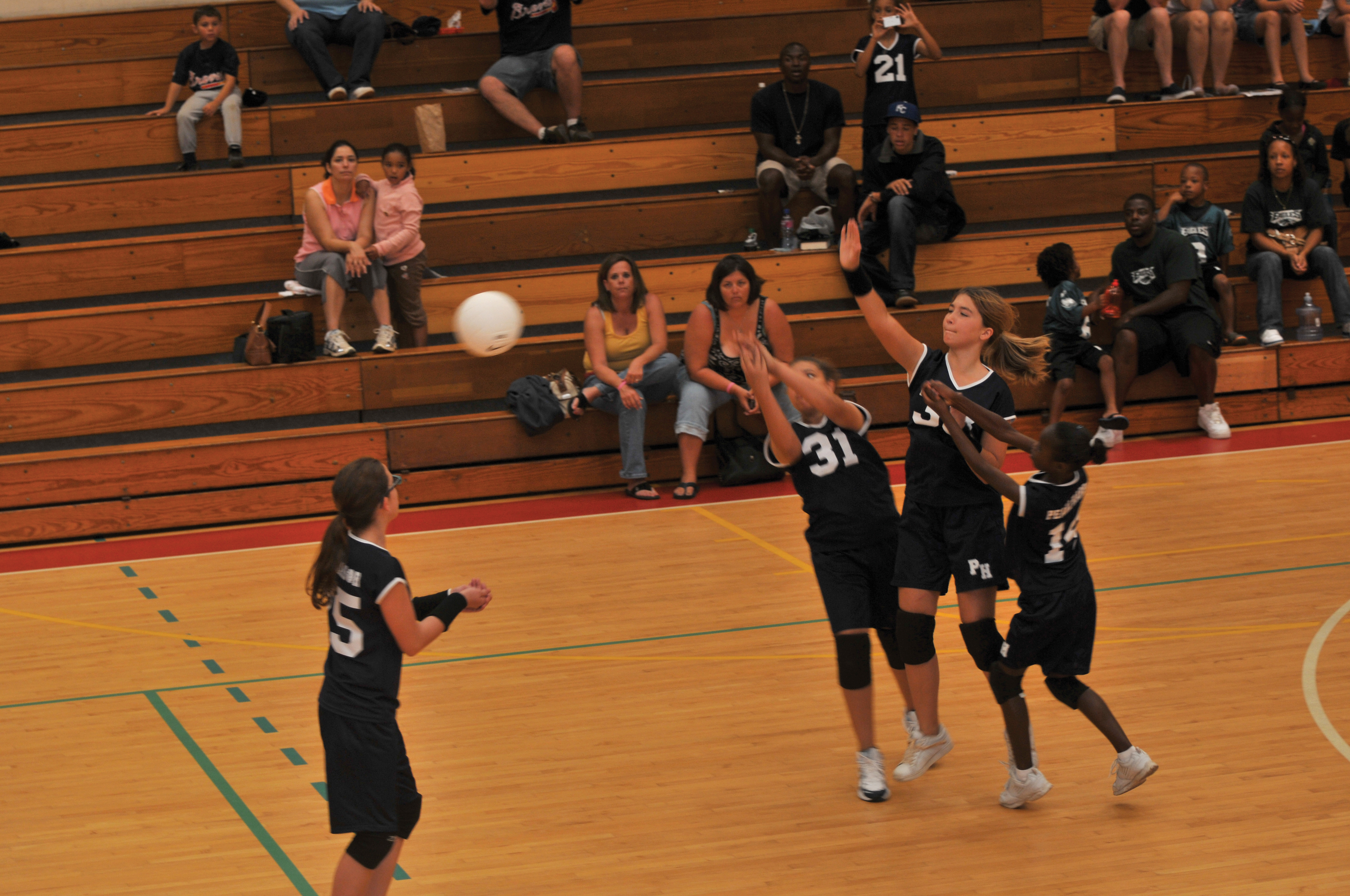 Girls in black uniforms playing volleyball in an indoor gymnasium with spectators in bleachers.