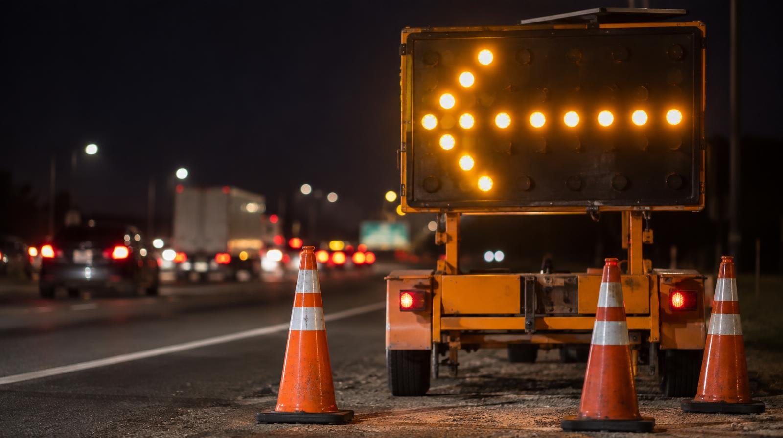 A yellow illuminated arrow board sign on a dark highway shoulder surrounded by orange traffic cones.