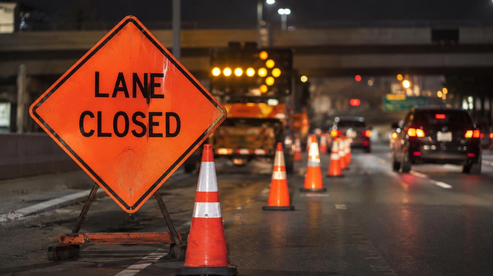 Orange signs and traffic cones narrowing a freeway to one lane at night.