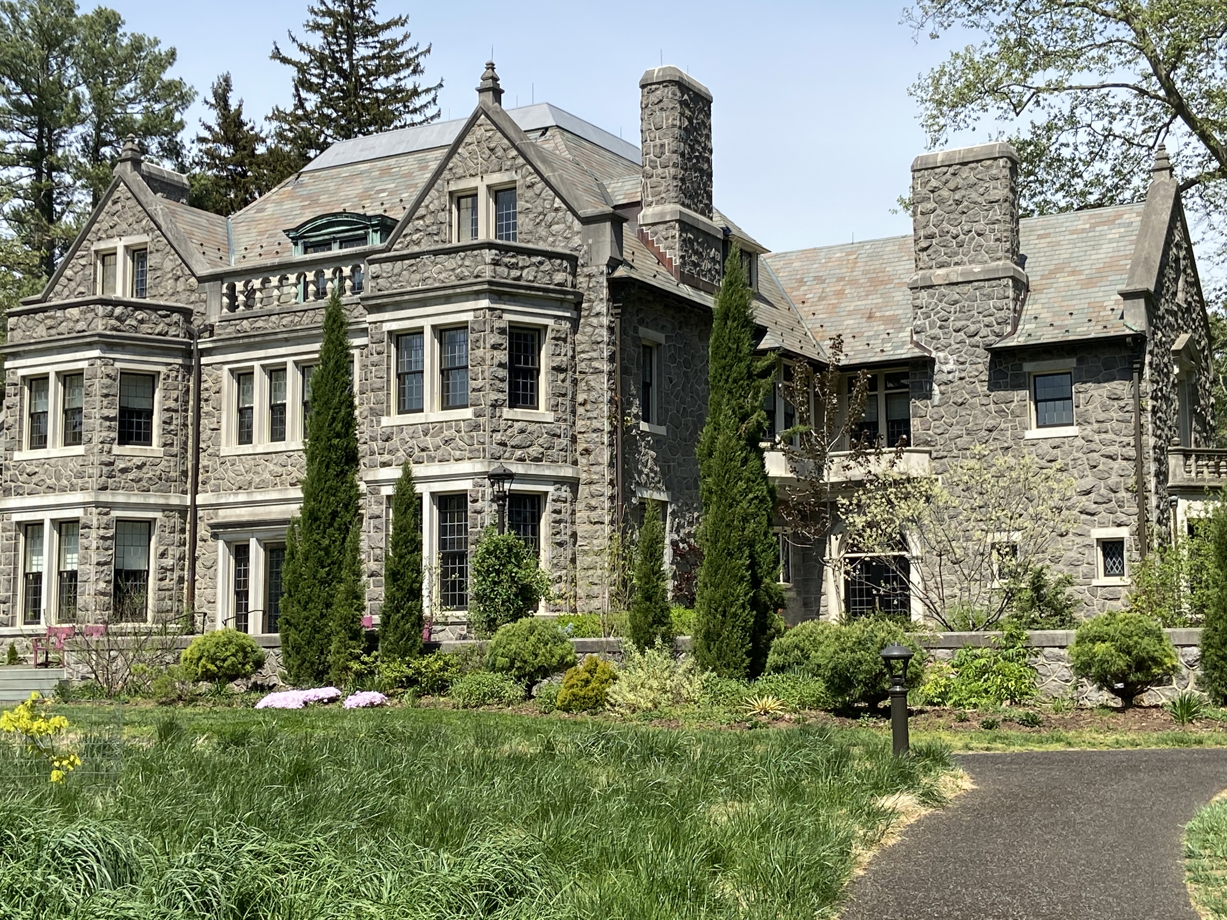 Large stone Tudor Revival mansion with tall evergreen shrubs on a manicured lawn