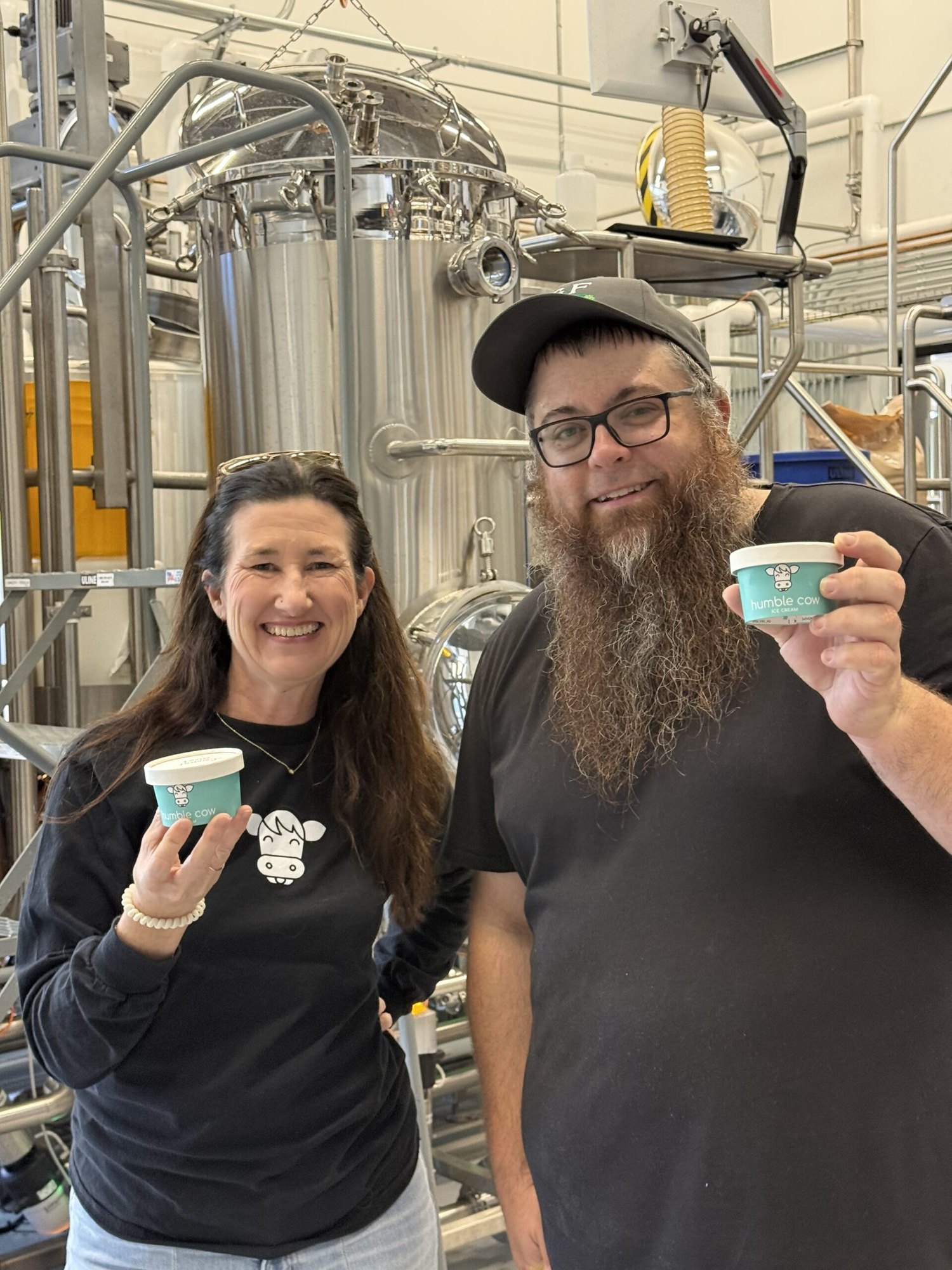Two people in black shirts hold small teal cups in front of large stainless steel brewing tanks.