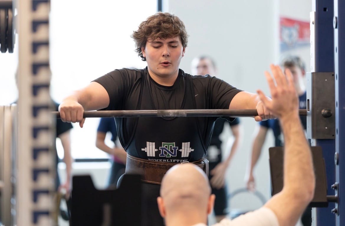 A young man in a black New Trier shirt squats a loaded barbell in a school weight room with a spotter in front.
