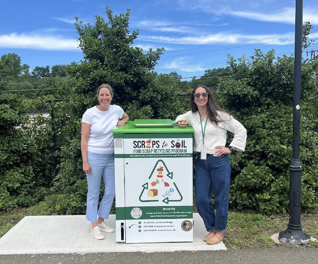 Two women standing beside a white Scraps to Soil food scrap recycling smart bin outdoors on a concrete pad.
