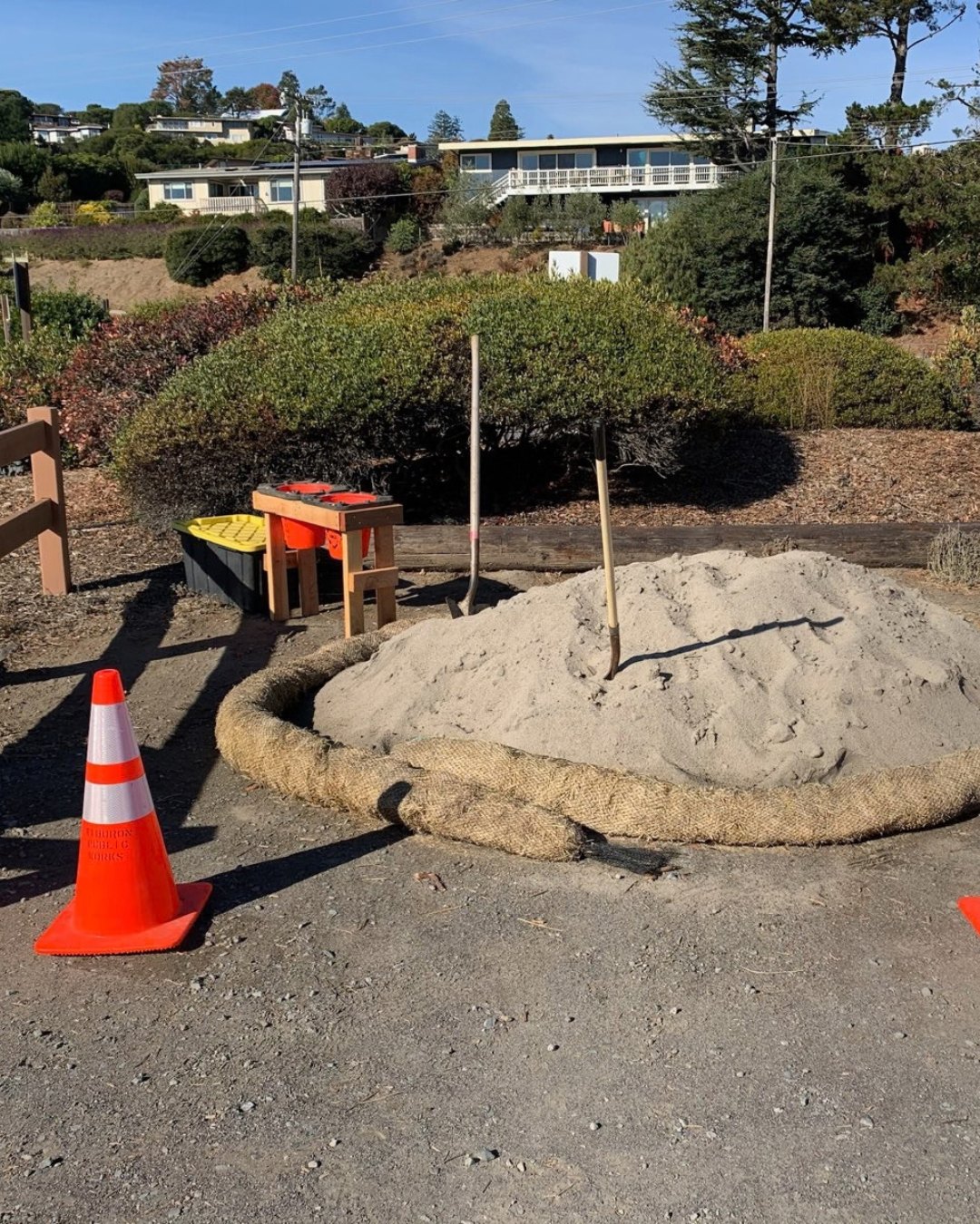 A mound of sand with sandbag tubes and shovels in a gravel parking lot.