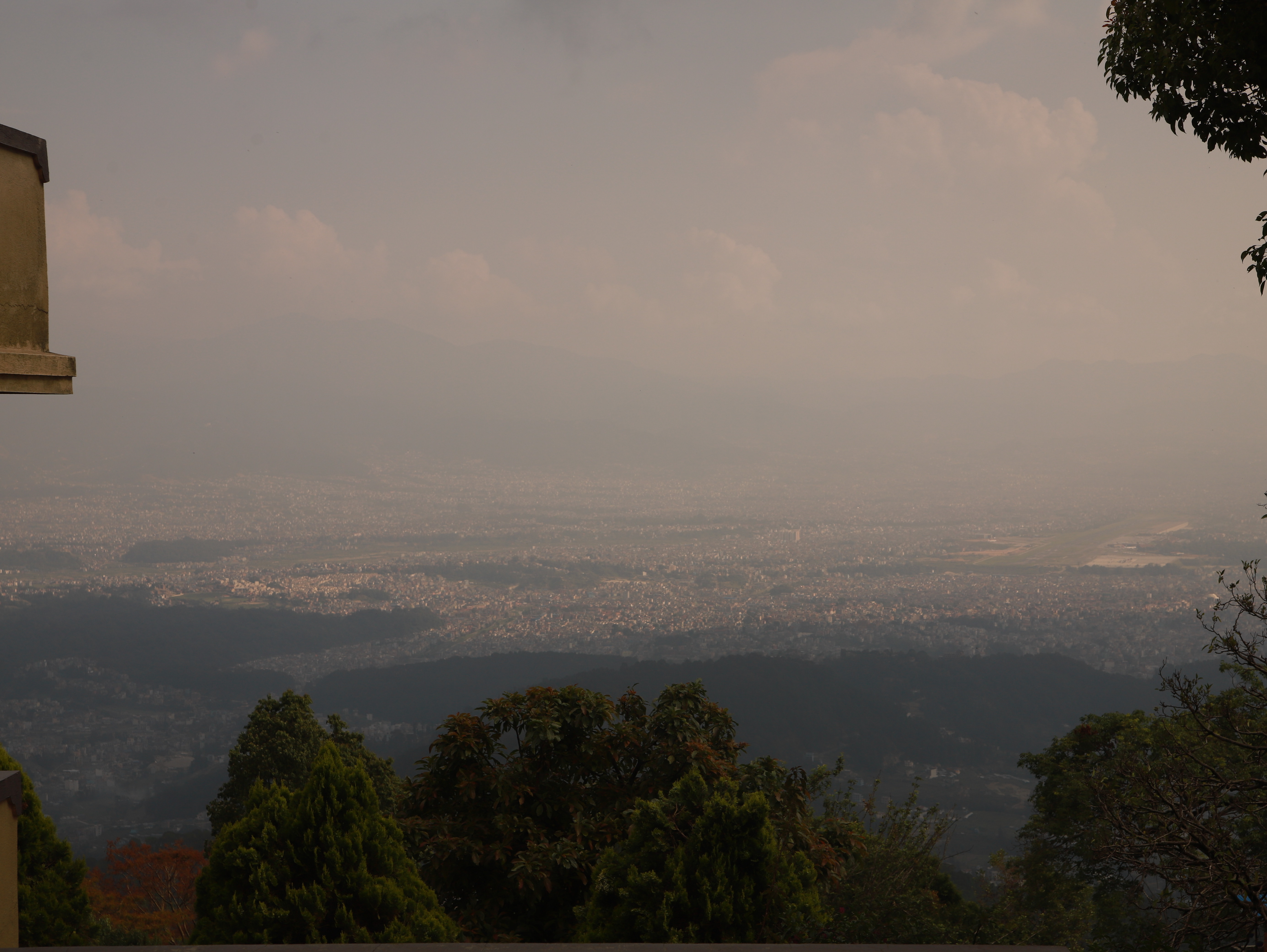 A city valley obscured by thick atmospheric haze, viewed from a forested hillside.
