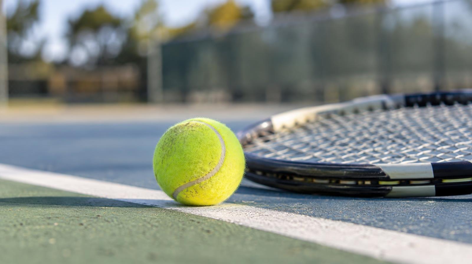Yellow tennis ball resting beside a racket on a hard court baseline in afternoon sunlight.