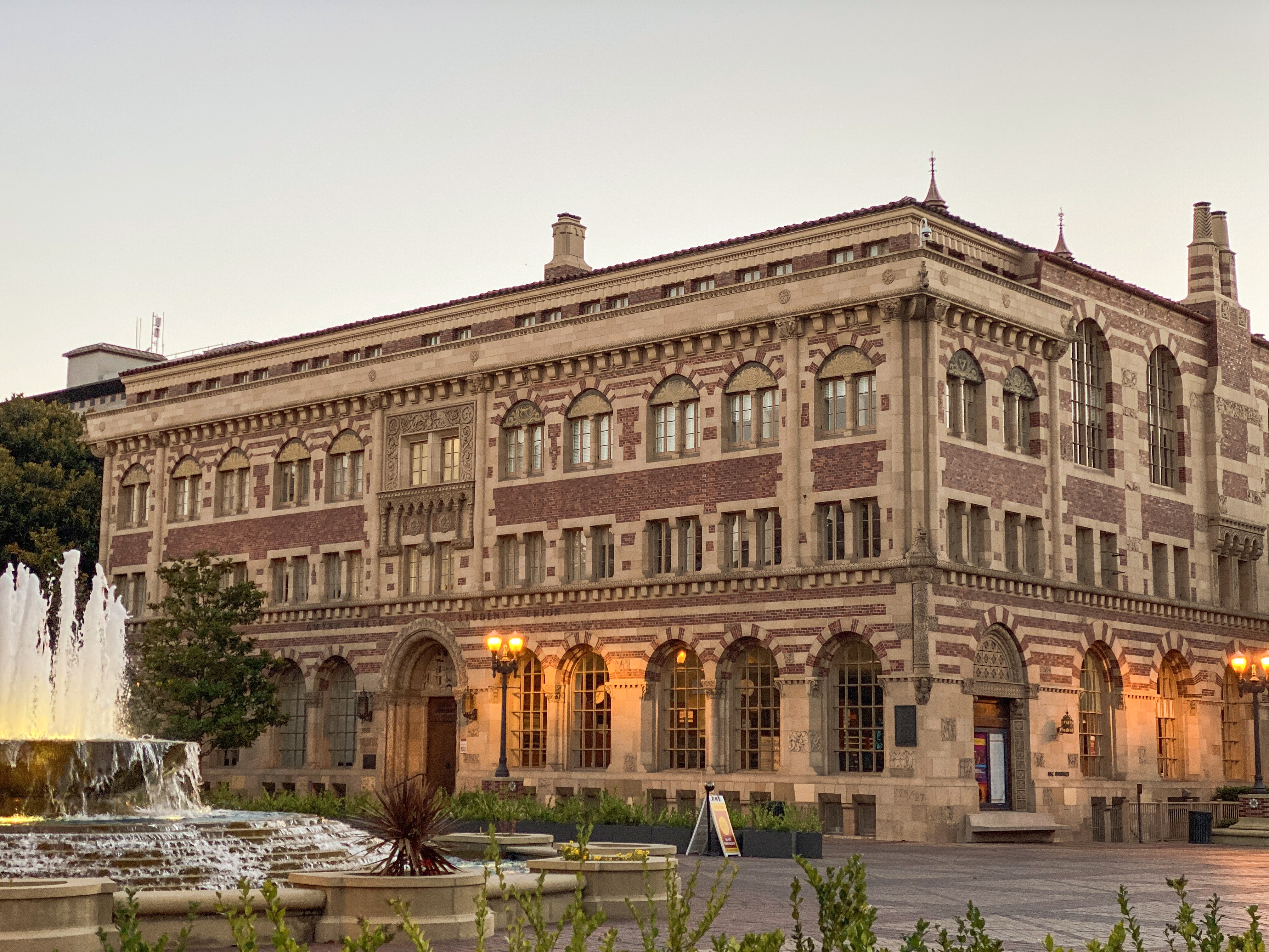 Ornate brick university building with arched windows and a fountain at dusk.