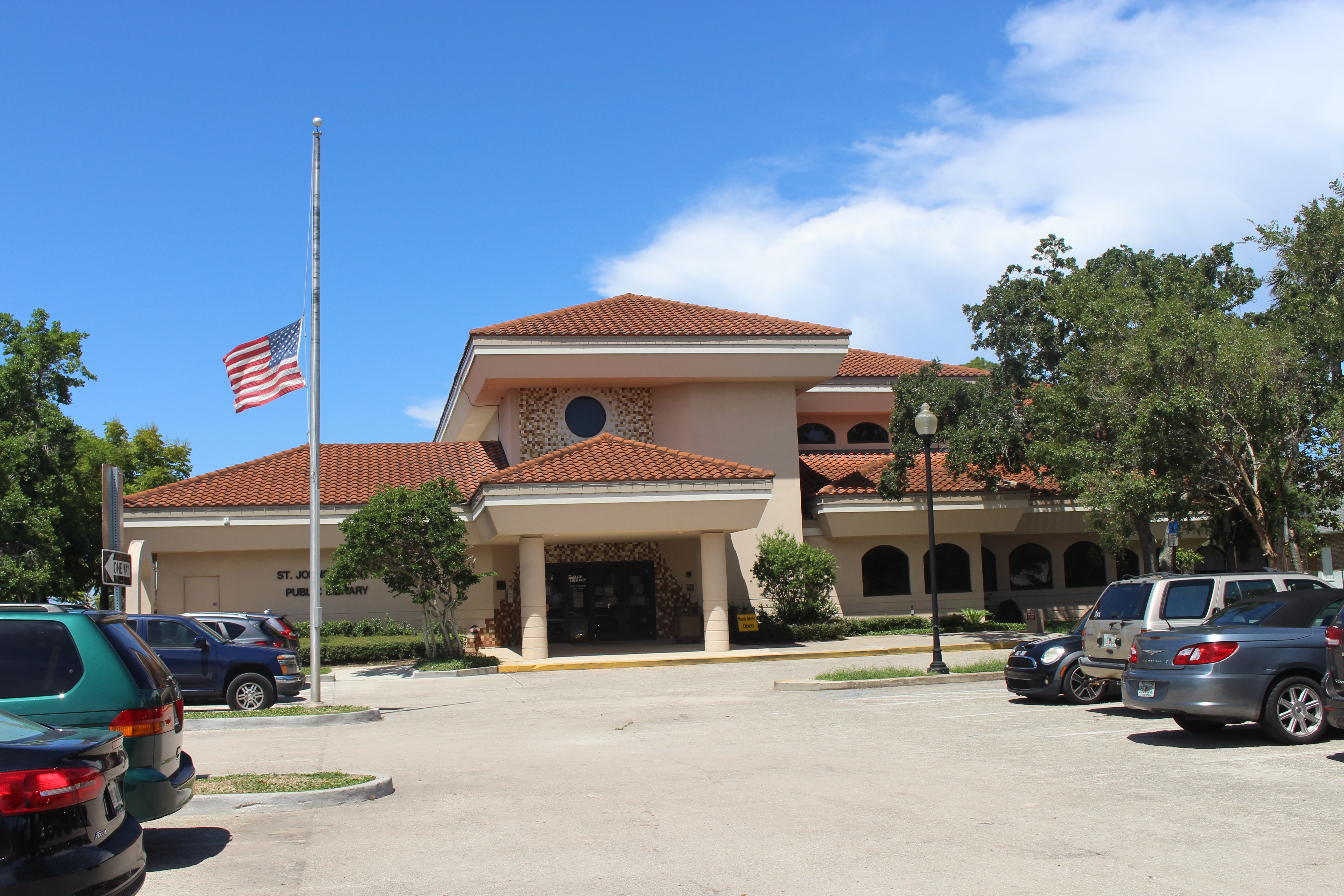 Two-story stucco library building with red tile roof and flagpole out front.