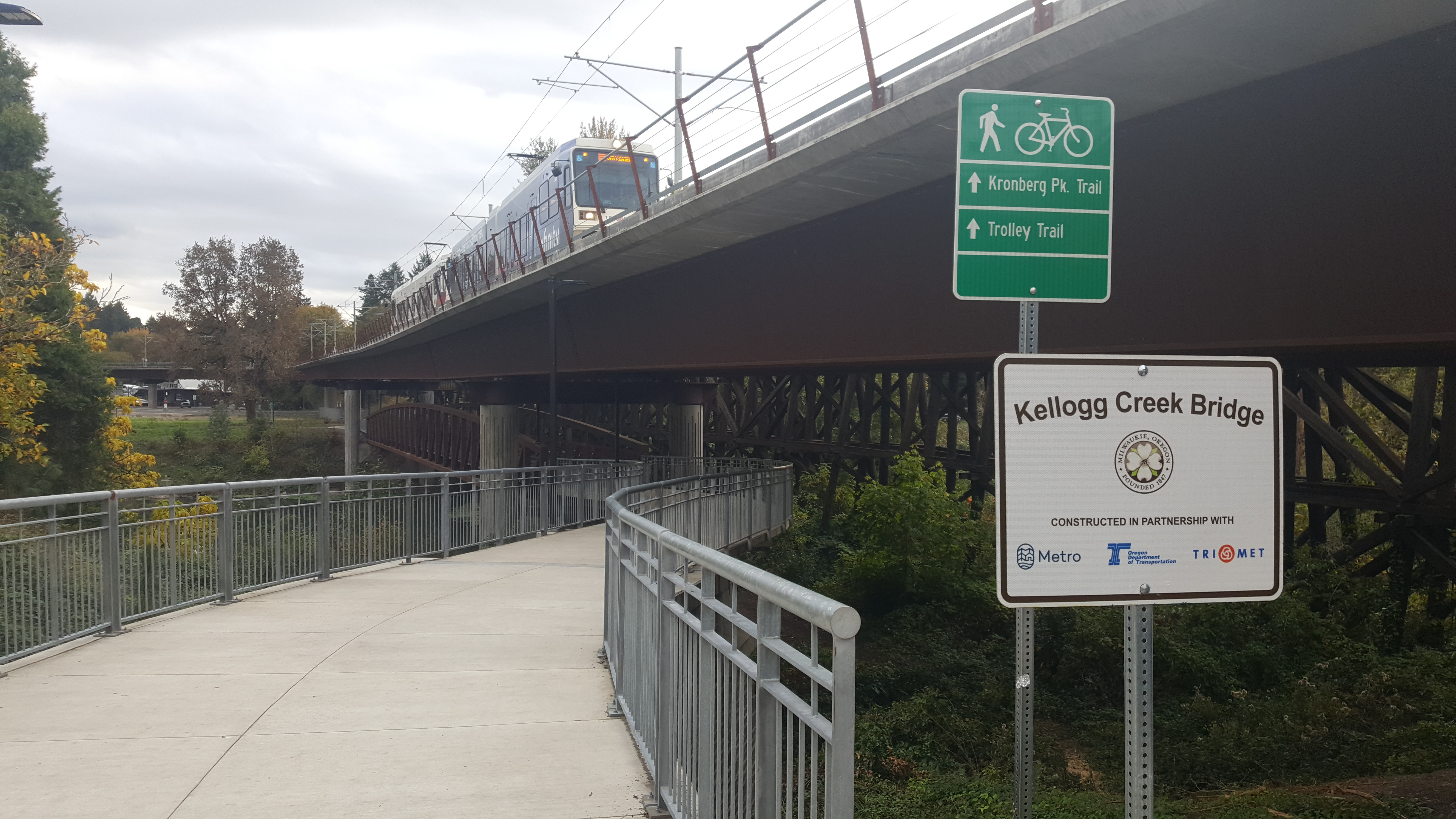 Pedestrian path with Kellogg Creek Bridge sign and light rail train crossing an elevated bridge in Milwaukie.