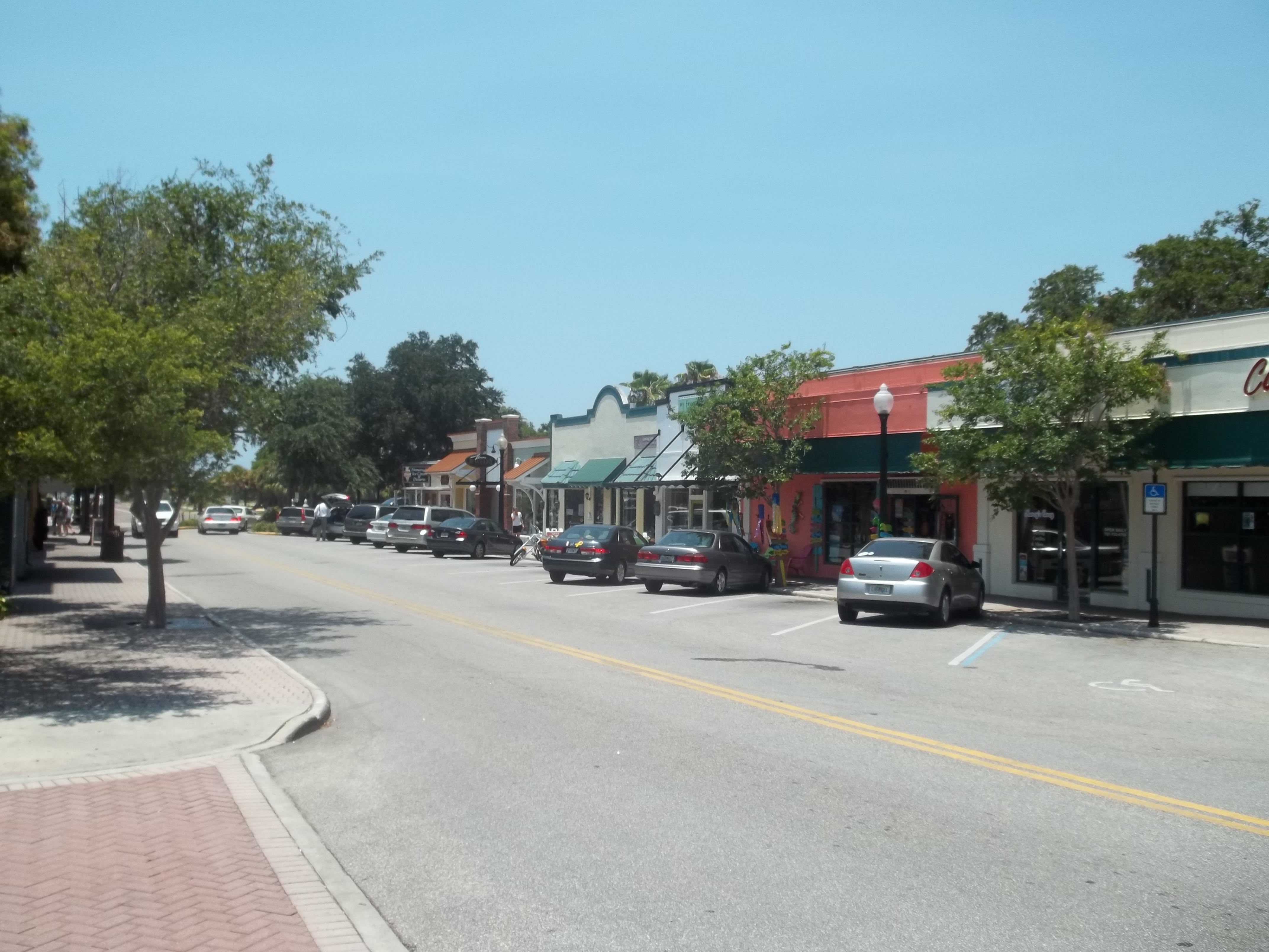 Tree-lined downtown street with low-rise commercial storefronts and parked cars on a sunny day.