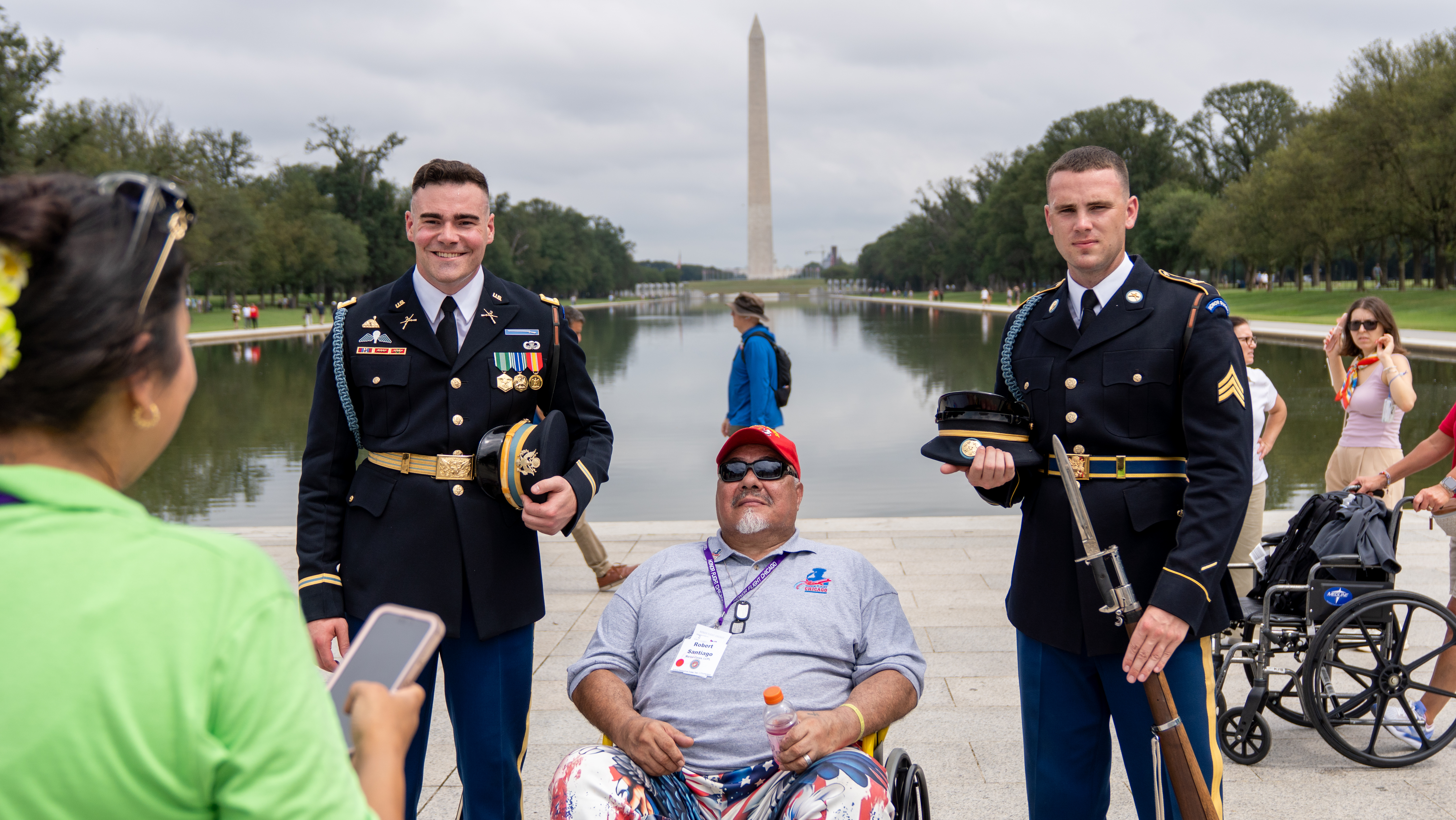 Two soldiers in dress uniforms flank a veteran in a wheelchair near a reflecting pool.