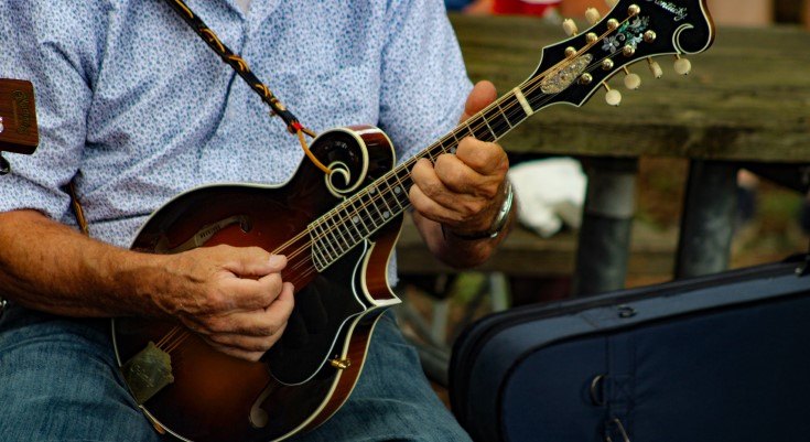 Person in a blue patterned shirt playing a sunburst mandolin outdoors.