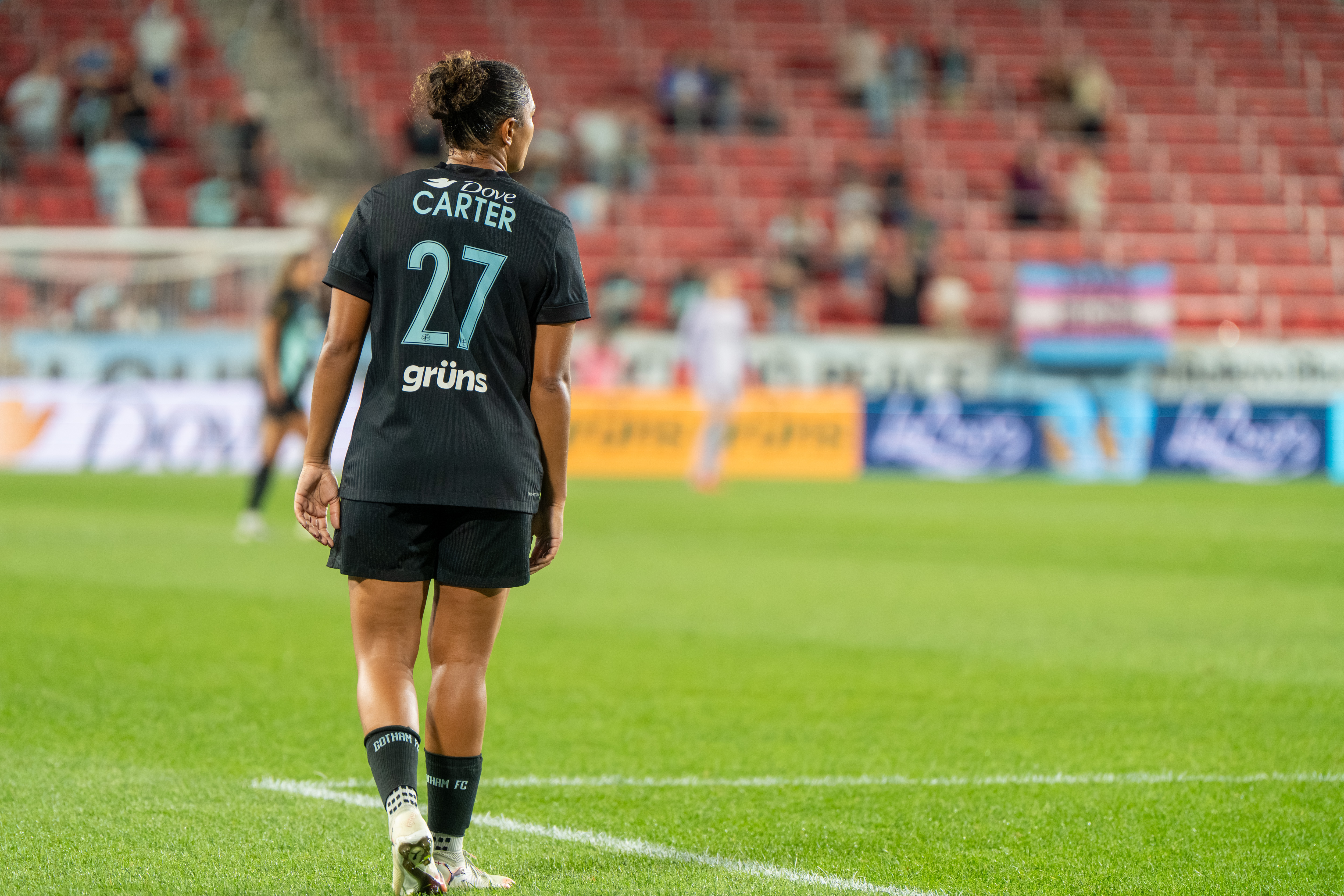 A soccer player in a black jersey numbered 27 stands on a pitch with stadium seating in the background.
