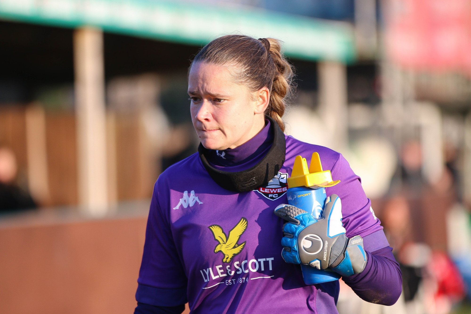 Female goalkeeper in purple Lewes FC kit wearing blue gloves and holding a water bottle.