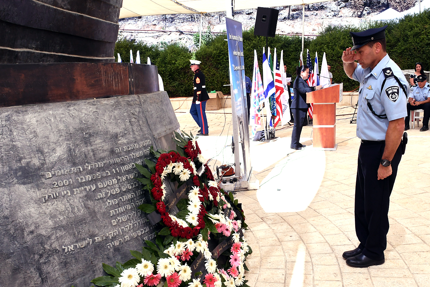 A uniformed officer salutes before a stone memorial with floral wreaths and flags in the background.