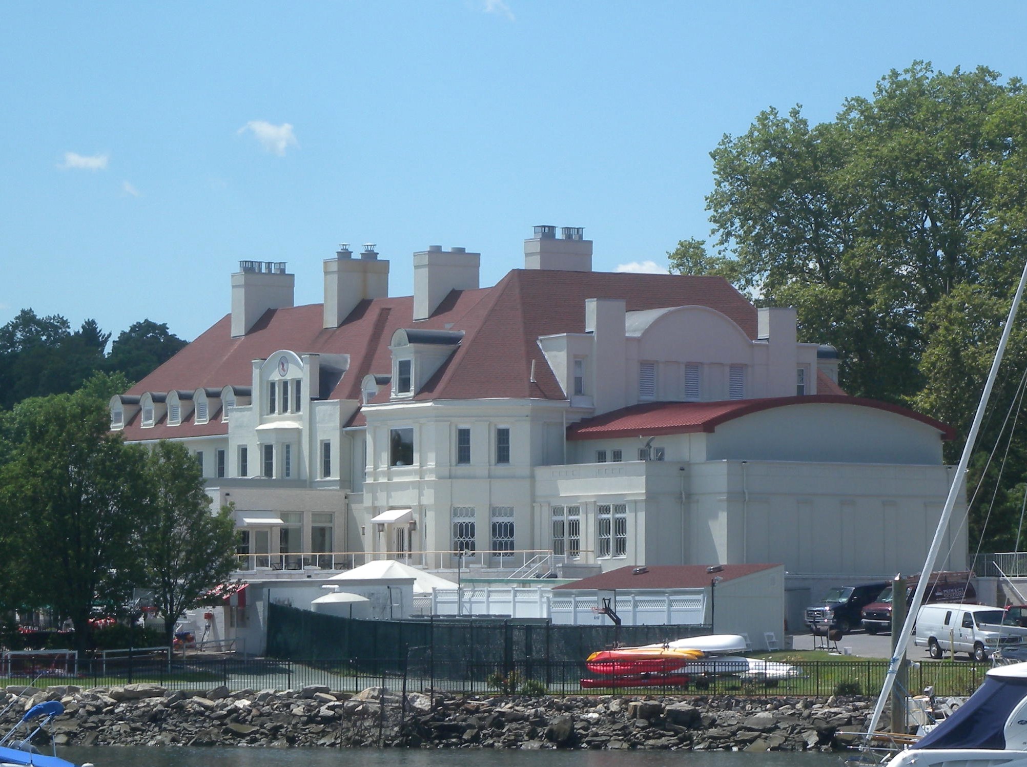 Large white waterfront clubhouse with red roofs viewed from the water on a sunny day.