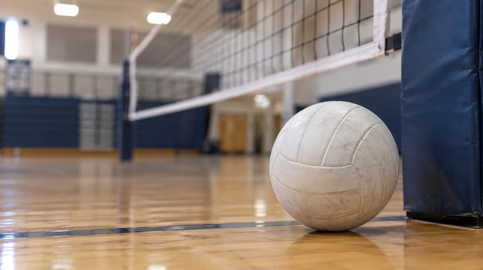 A white volleyball on a hardwood gym floor next to a volleyball net.