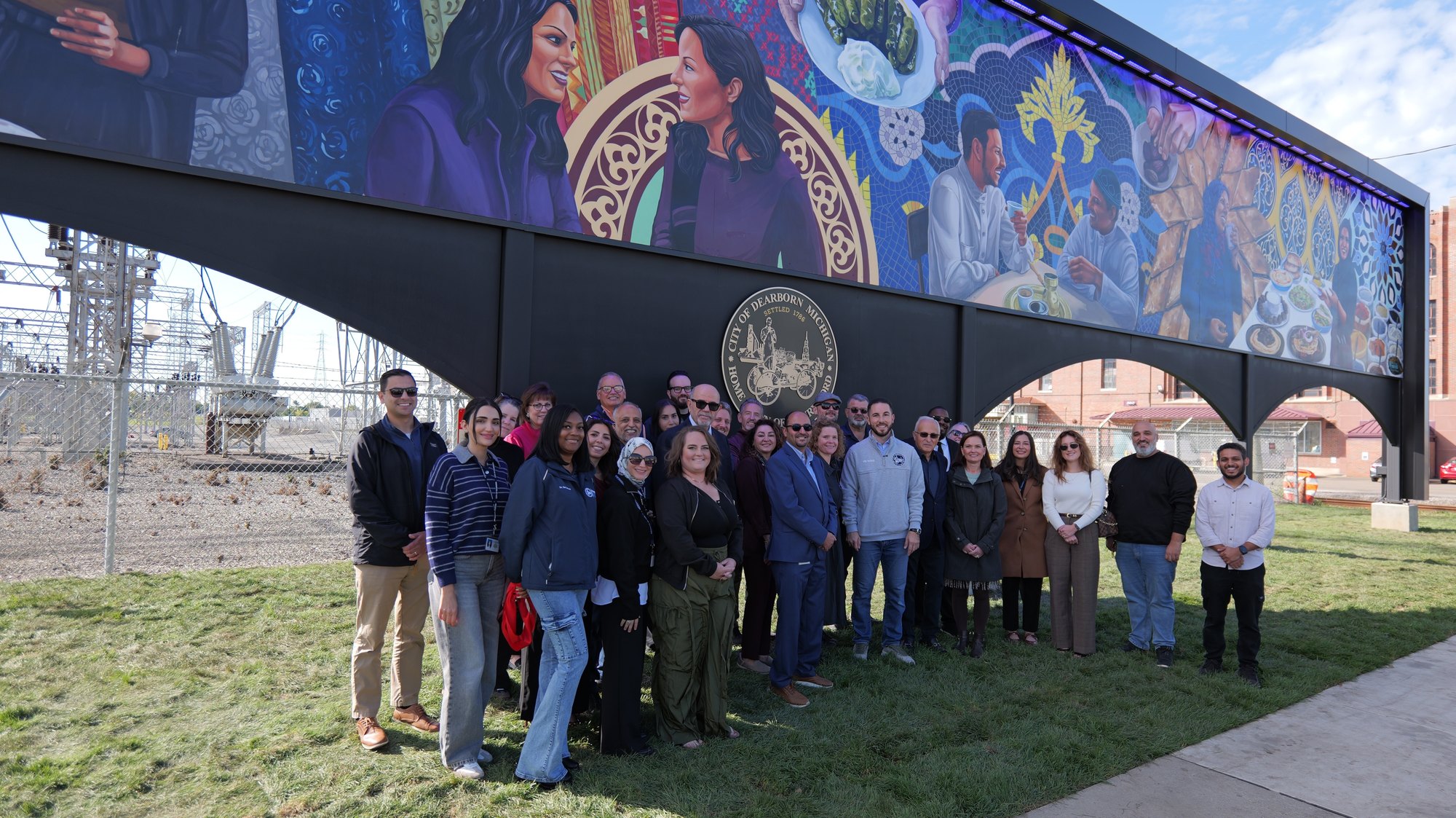 Large group of people standing in front of a colorful large-scale mural on a metal structure outdoors.