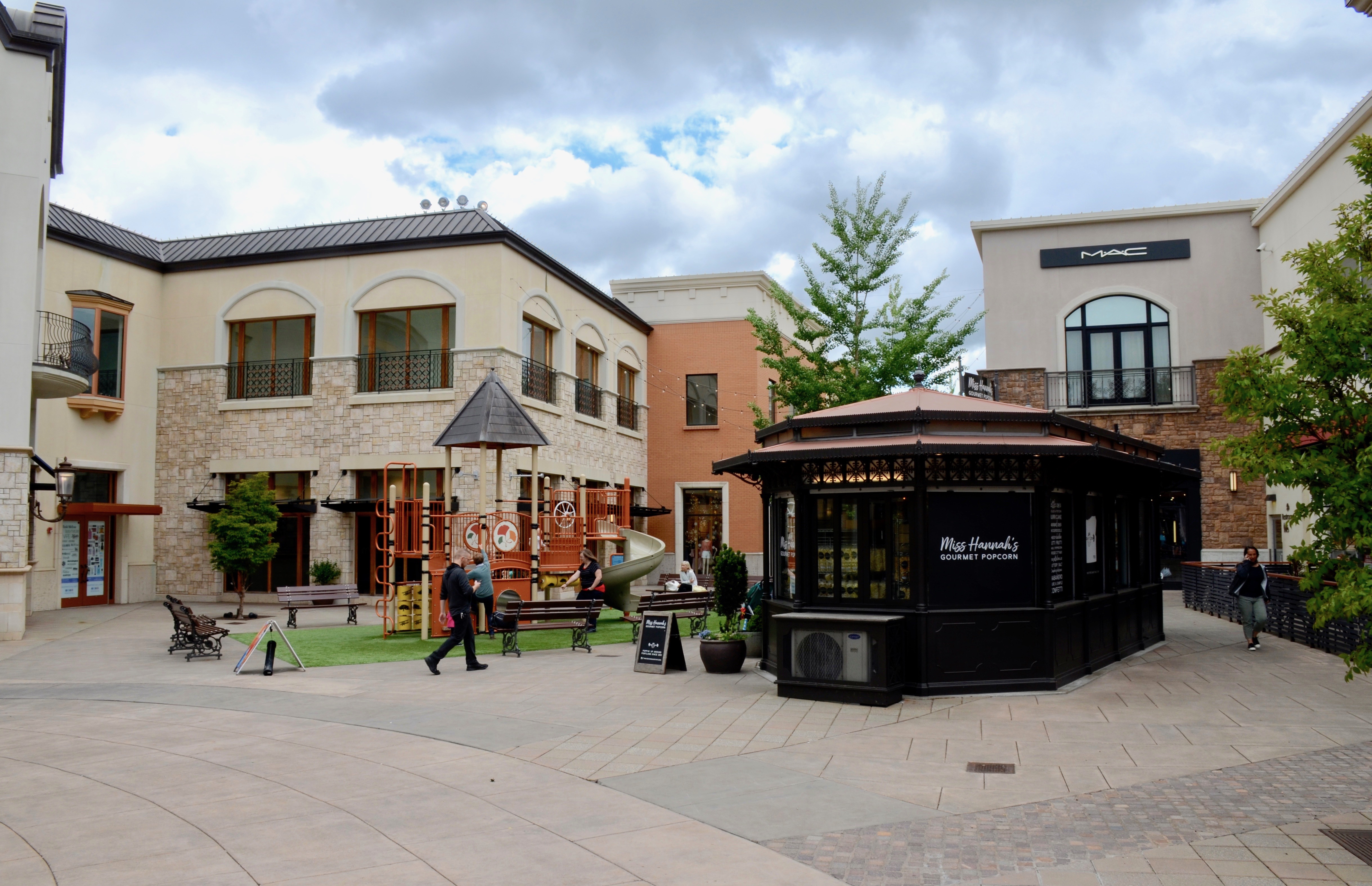 Outdoor shopping mall plaza with a central kiosk, children's play area, and retail storefronts.