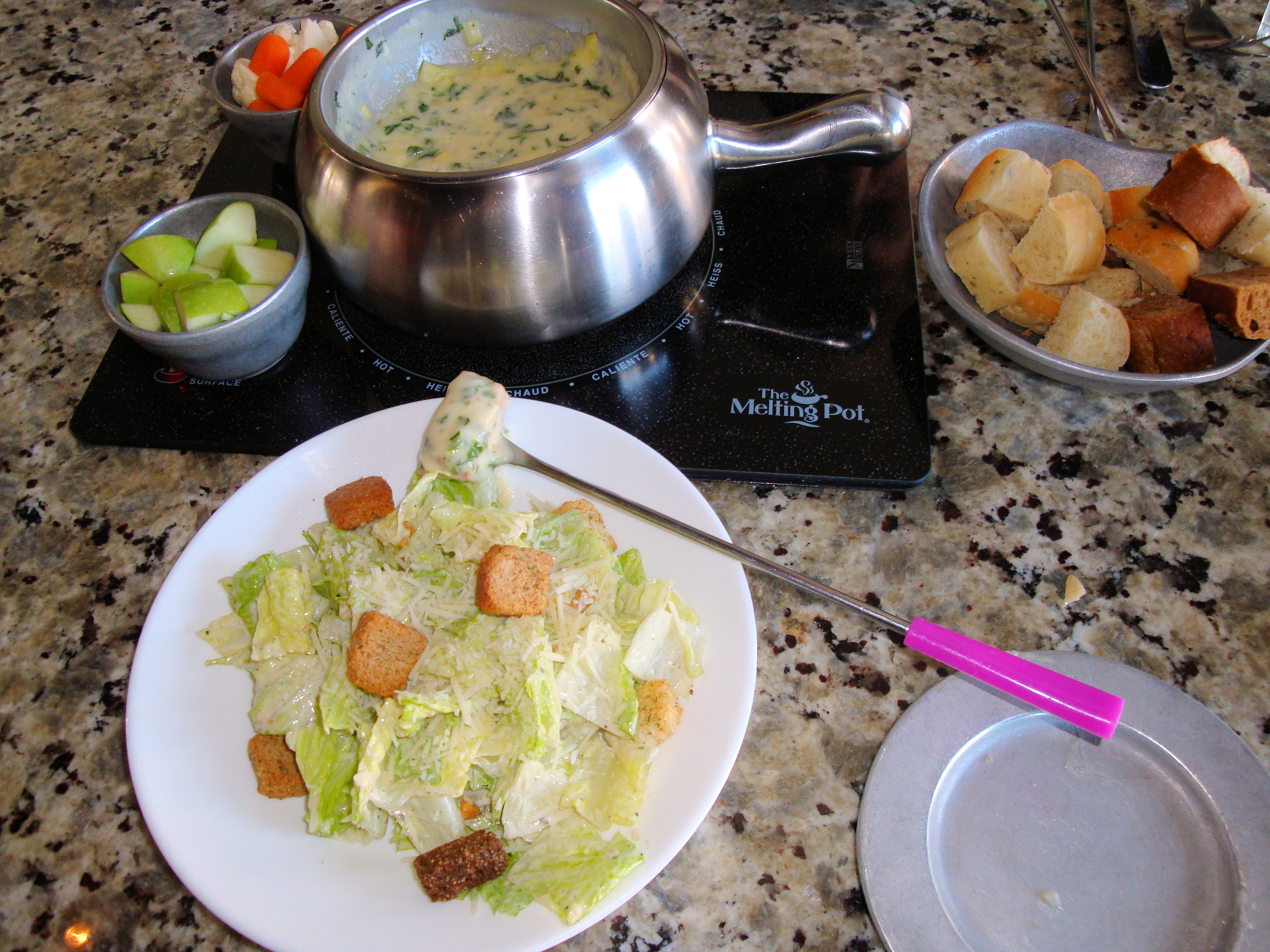 Restaurant table with a fondue pot, Caesar salad, dipping items, and colored fondue forks.