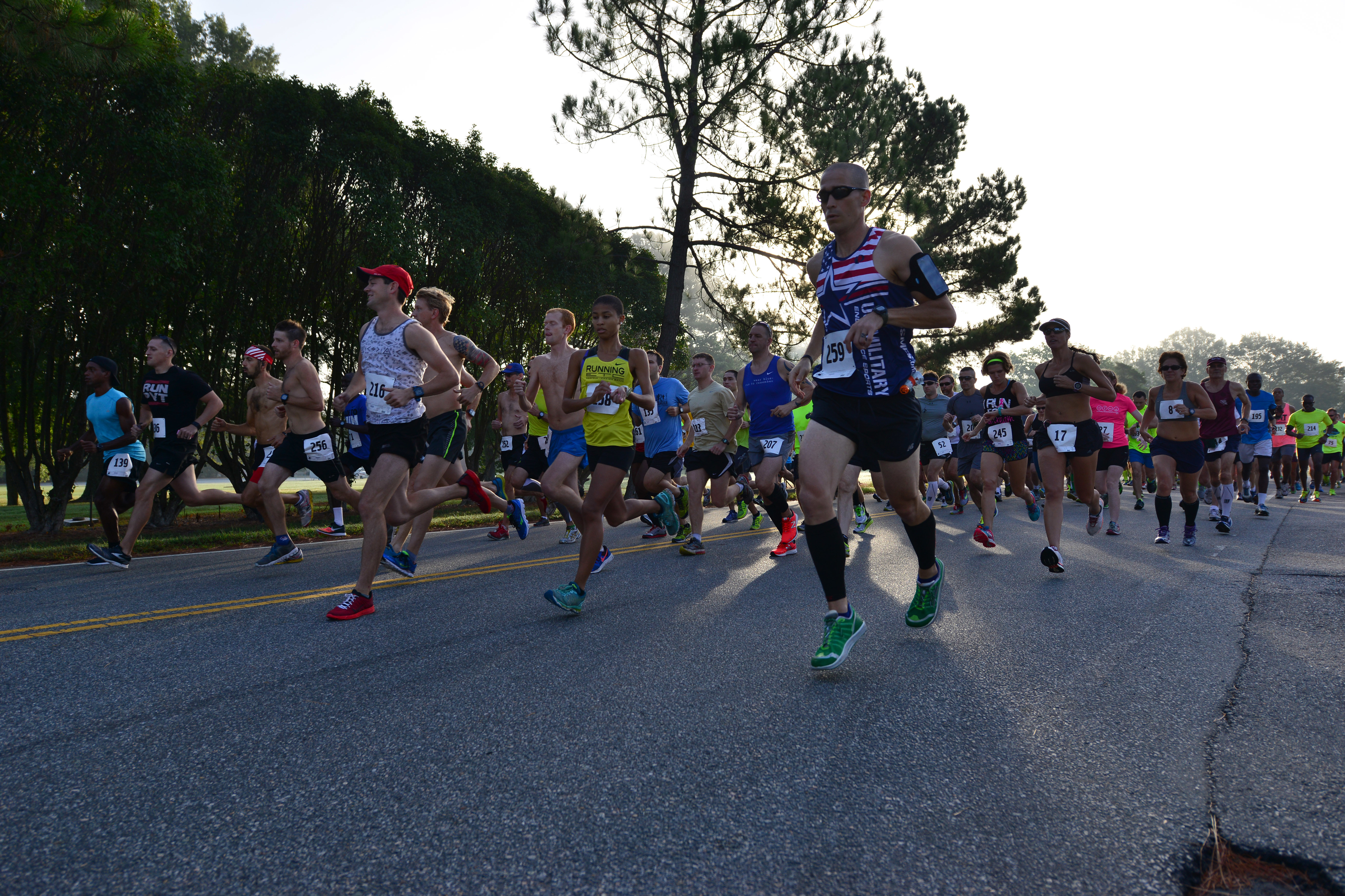 Large crowd of runners in race bibs launching from a starting line on a tree-lined road.