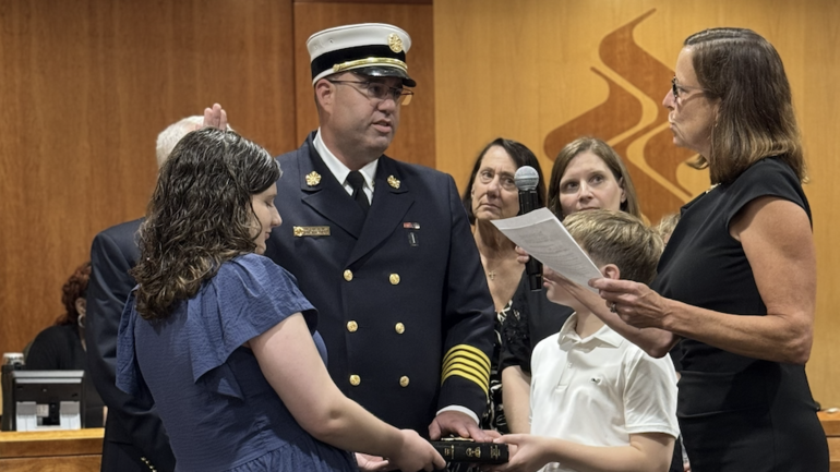 A man in a fire chief's dress uniform being sworn in by a woman reading from a document, surrounded by family members.