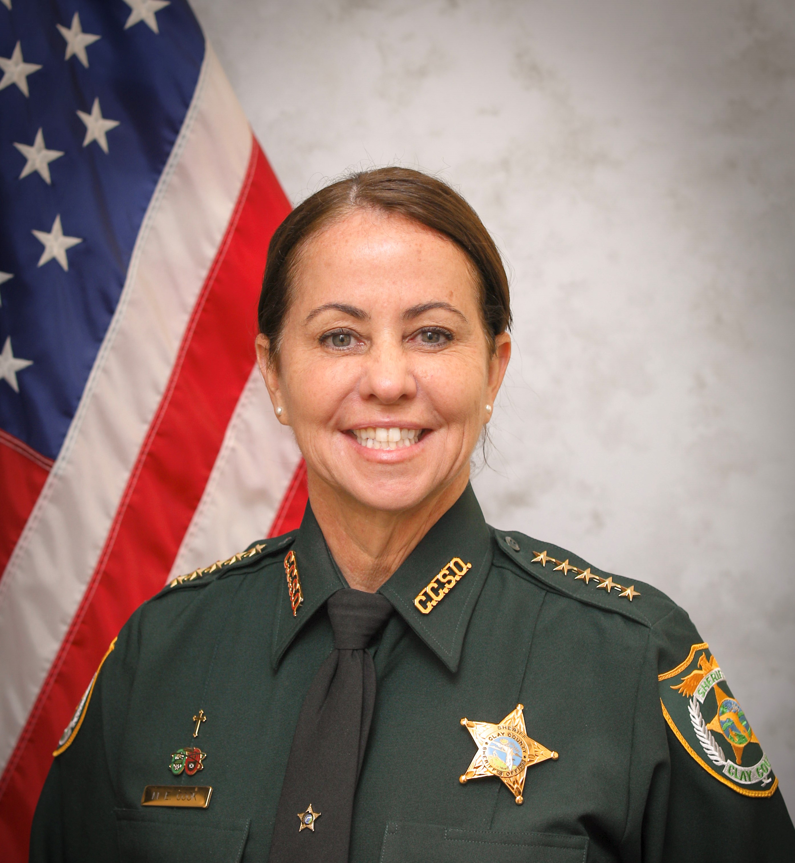 Woman in dark green sheriff's uniform with gold badge posed before an American flag.