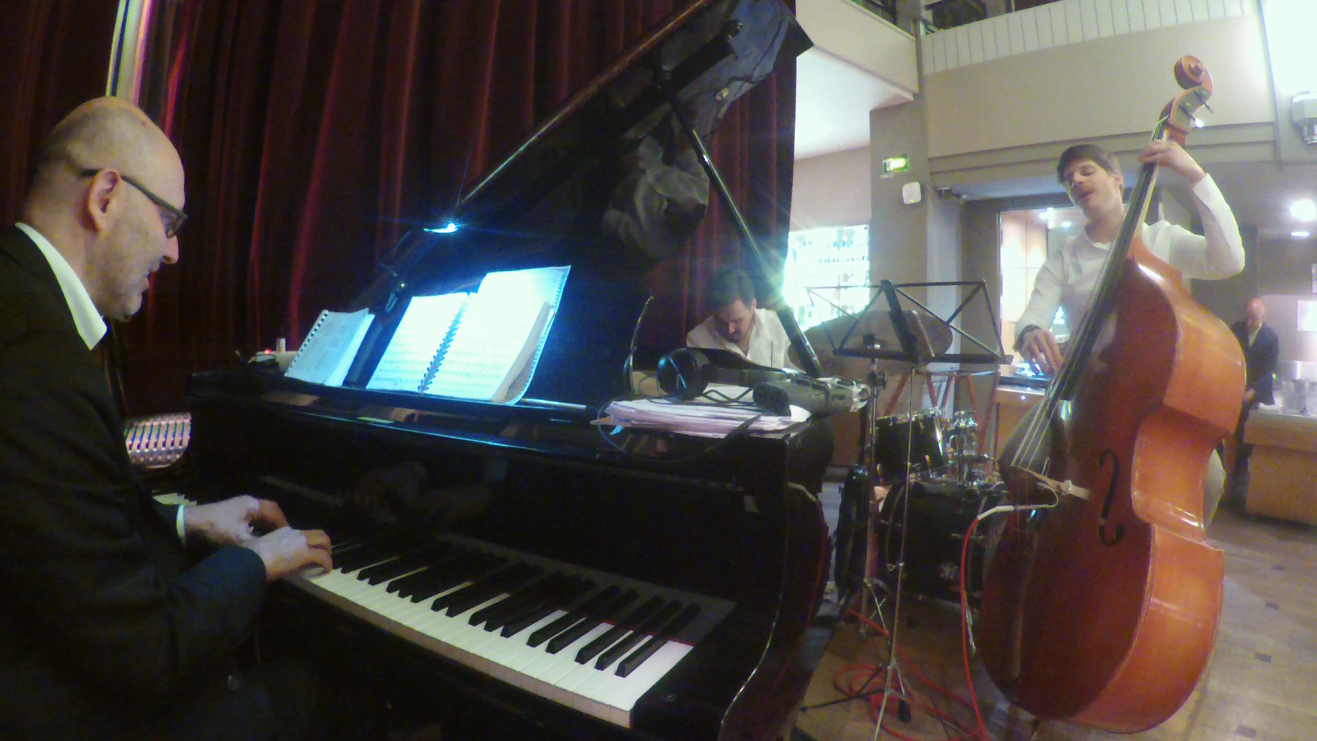 A pianist at a grand piano performs with a bassist and drummer in a warmly lit indoor venue.