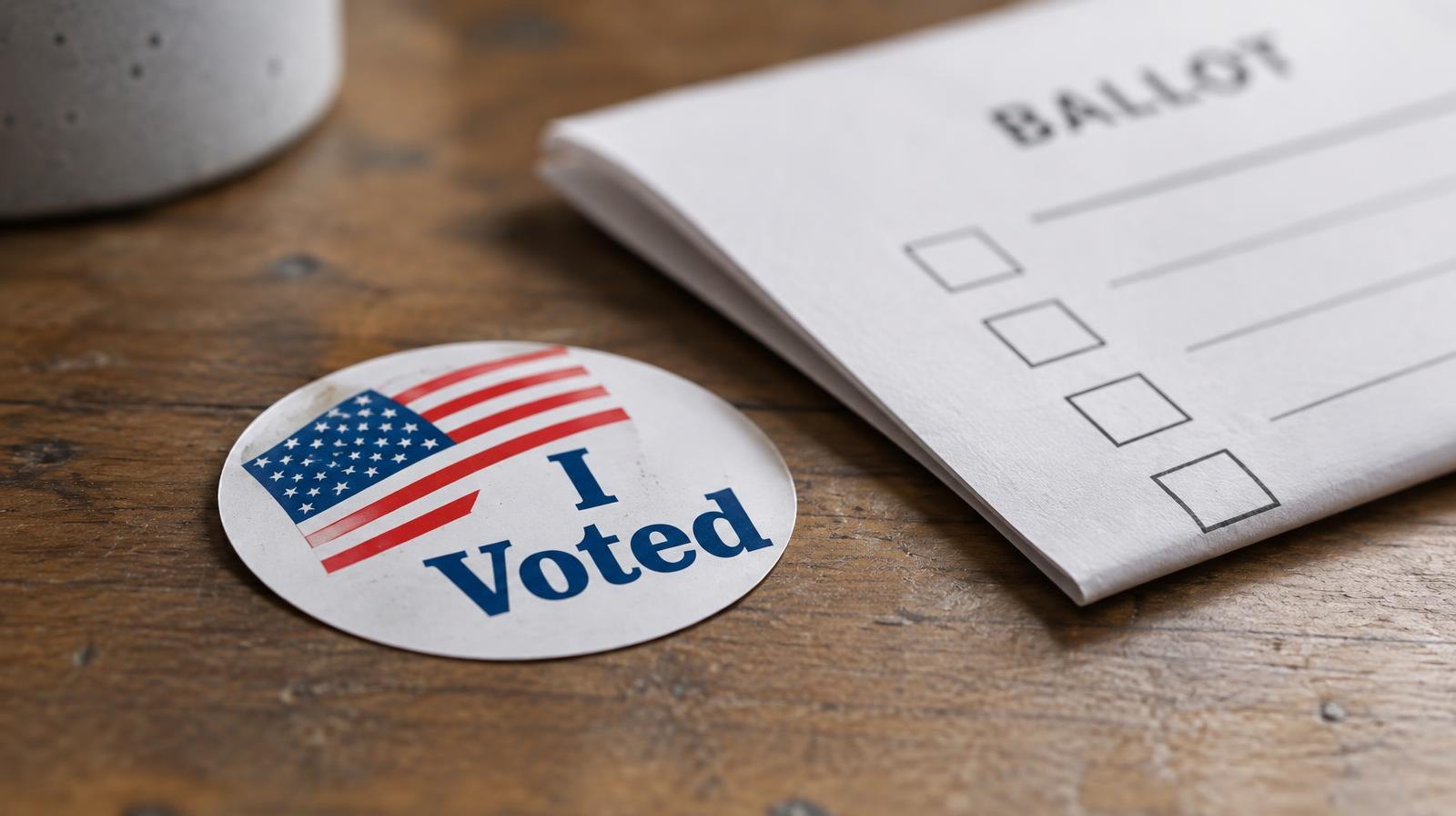 An I Voted sticker resting on a wooden surface beside a folded paper ballot.