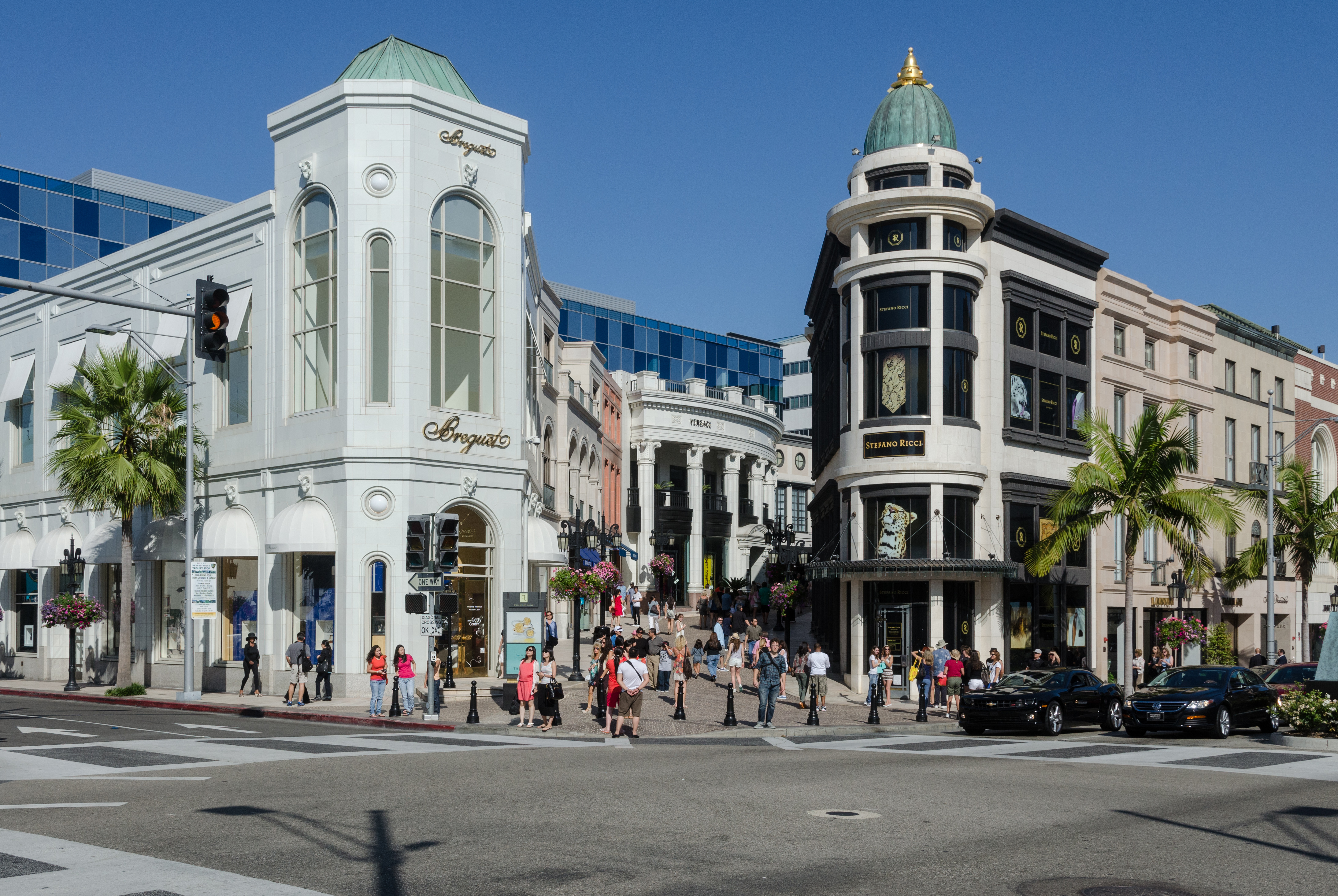 Street-level view of luxury retail buildings on Rodeo Drive with pedestrians crossing the intersection under blue skies.