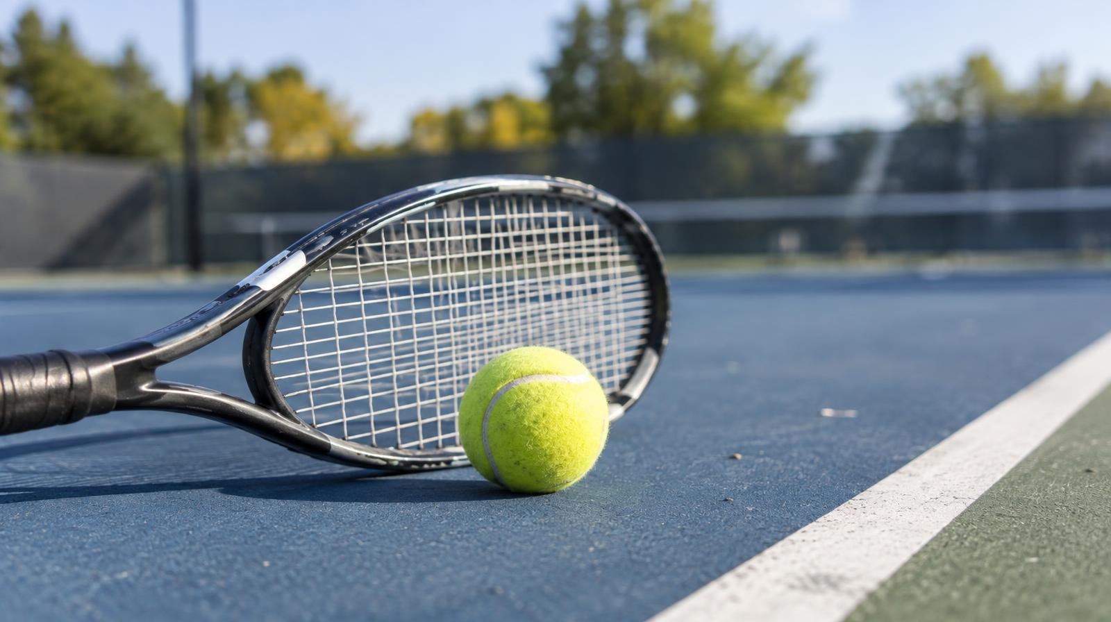 A tennis racket and yellow ball resting on a blue hard court surface.