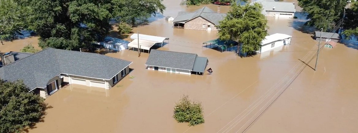 Aerial view of houses partially submerged in brown floodwater with utility poles visible.